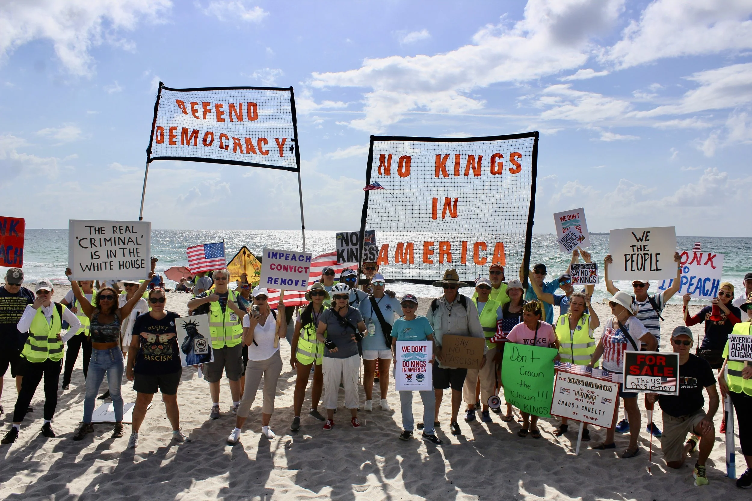 A large group of people participating in a protest on a beach, holding various signs and banners with messages against monarchy, democracy, and political issues, with flags and a partly cloudy sky in the background.