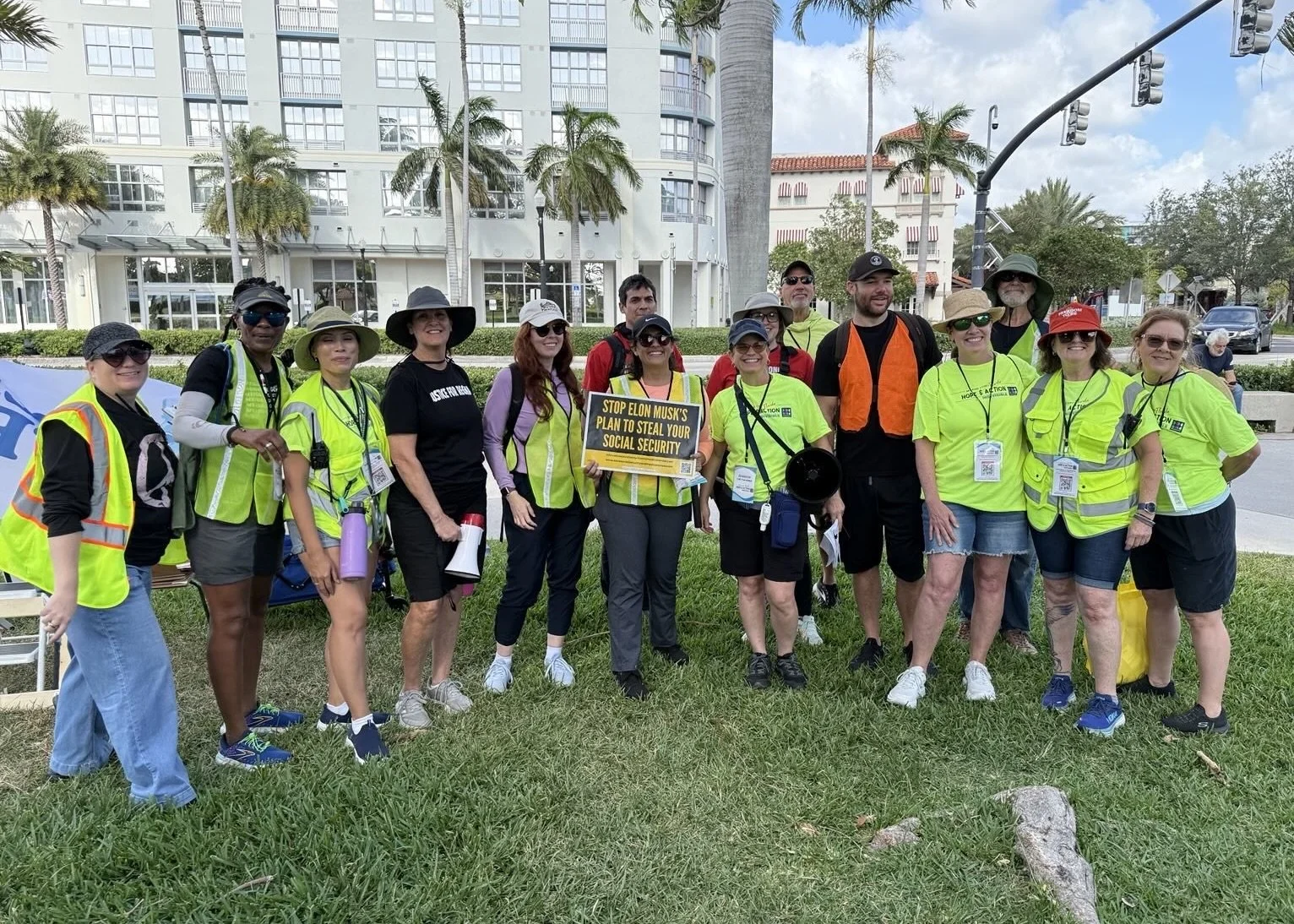 Group of people wearing yellow vests and hats, standing together outdoors, holding signs, in front of a building with palm trees.