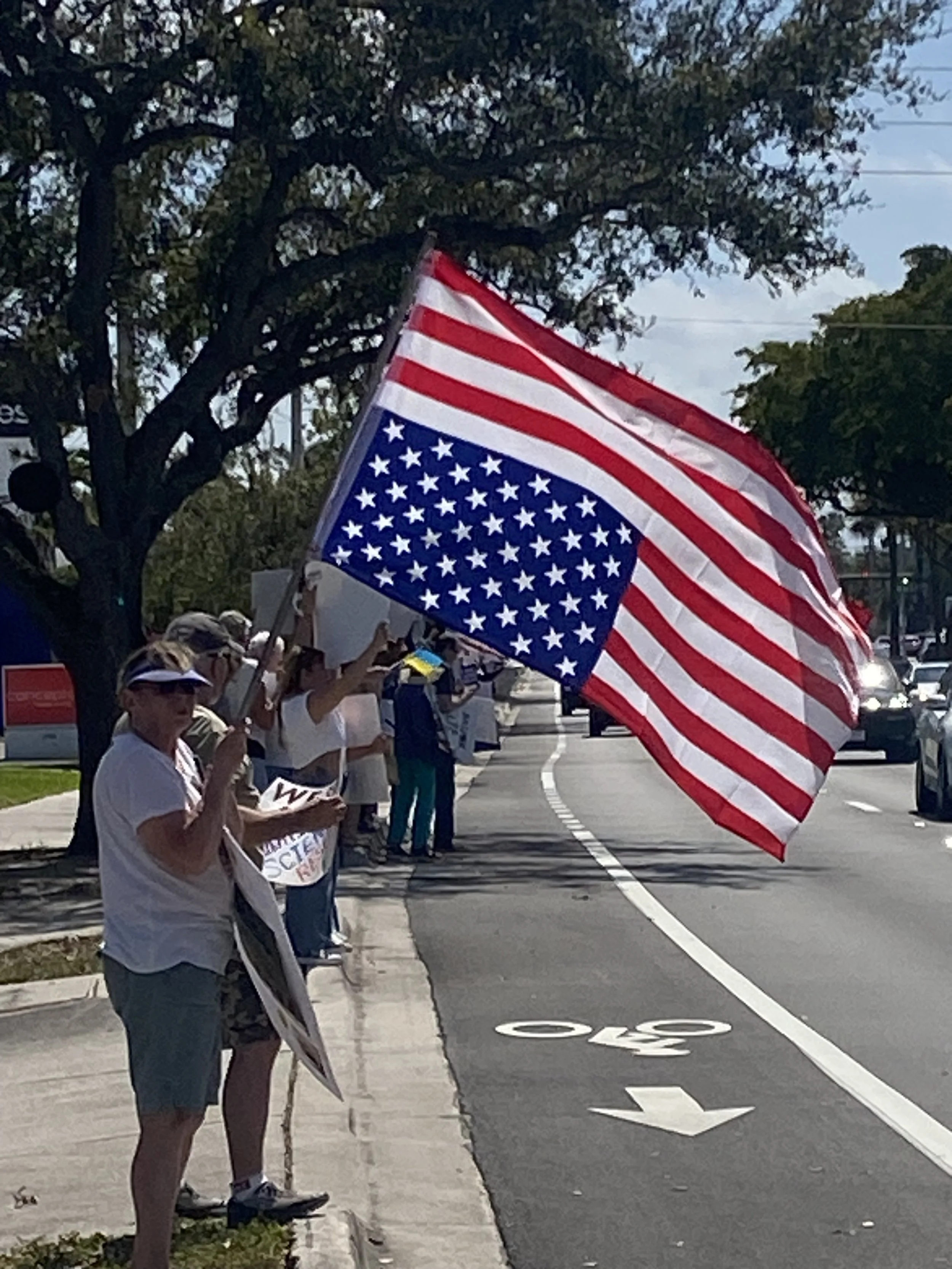 People participating in a parade or rally outdoors, holding an American flag. Some are wearing hats and sunglasses, and a woman is holding a sign.