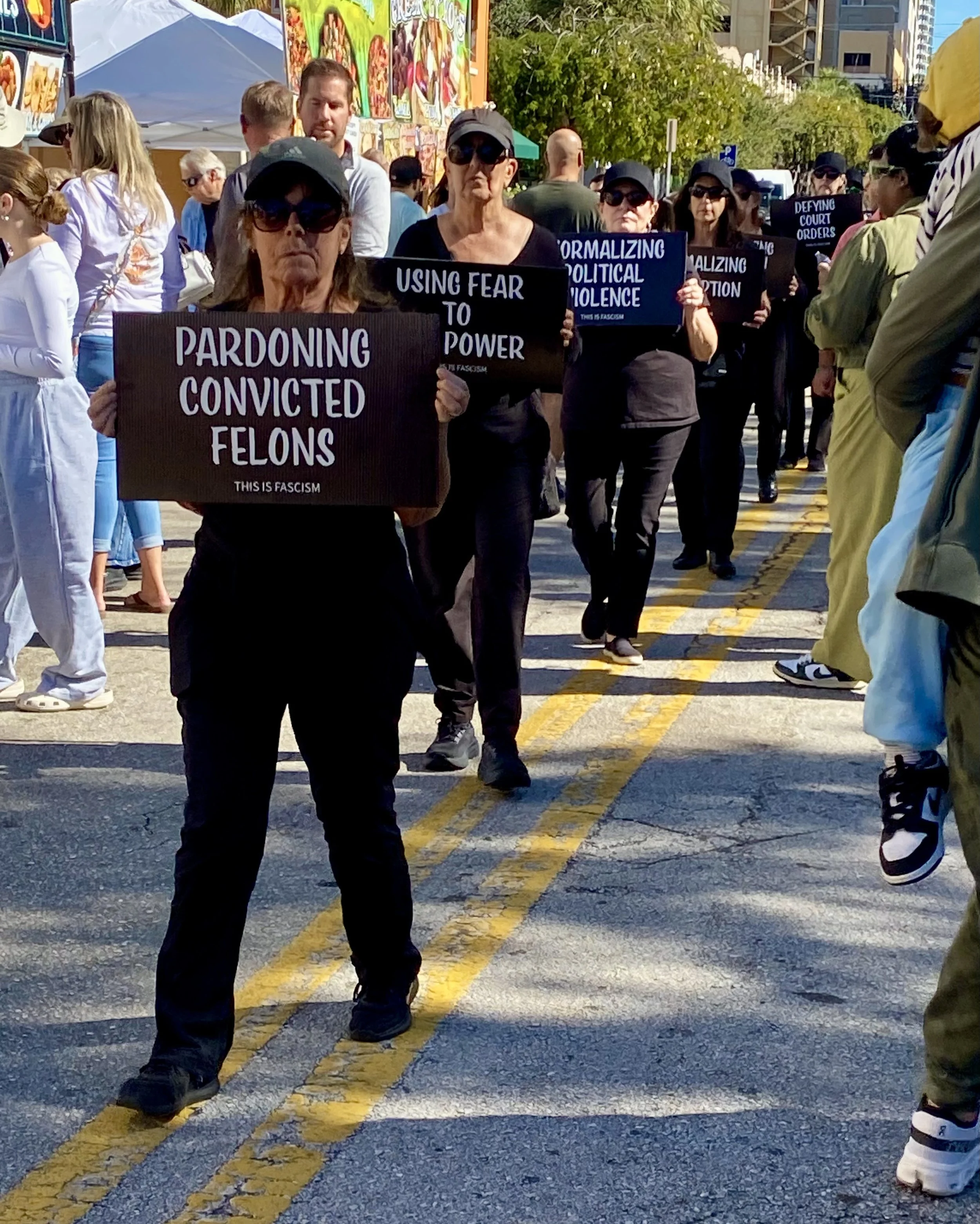 Protesters marching on a city street holding signs with messages about criminal justice and political issues, wearing black clothing and sunglasses.