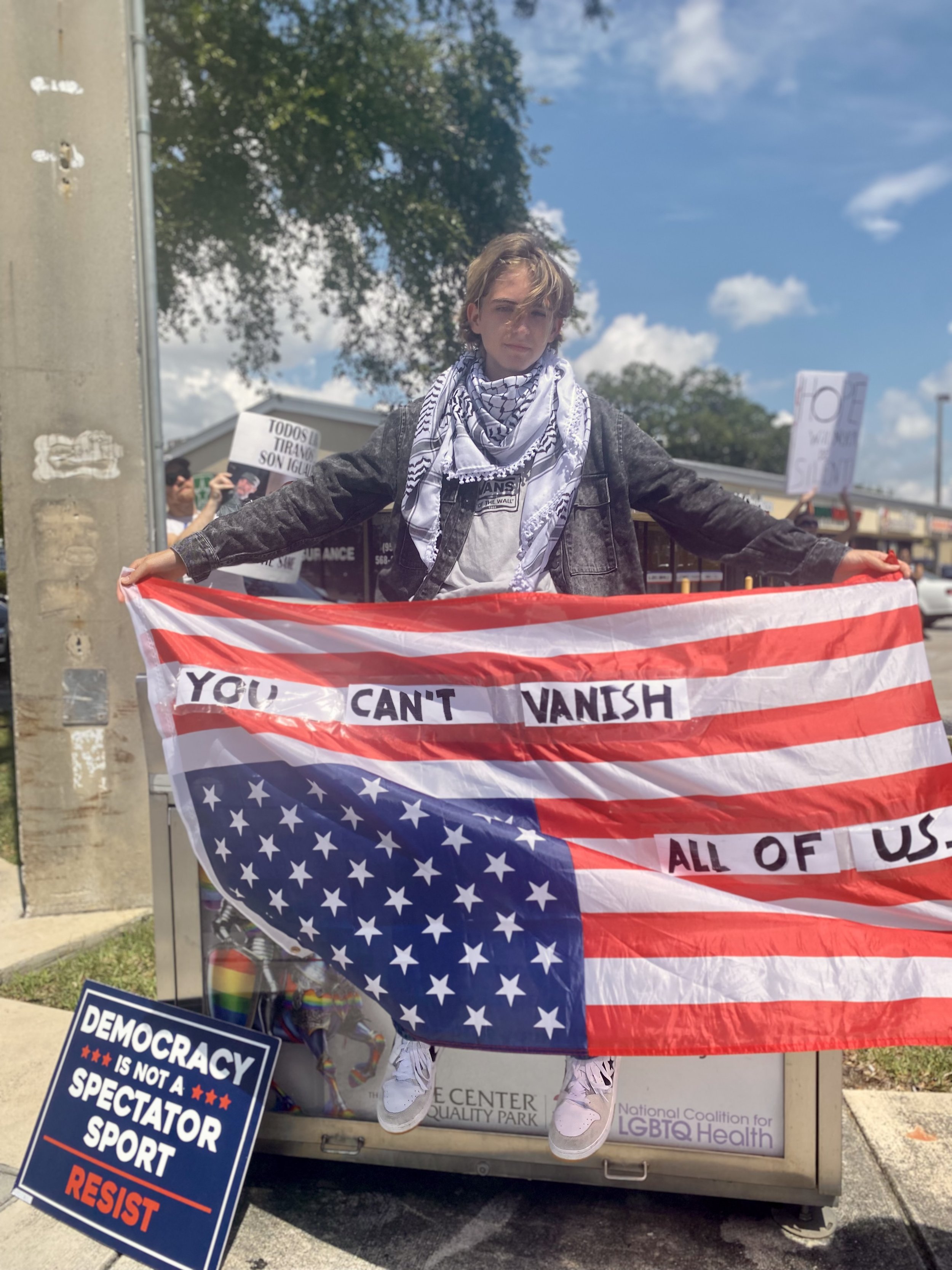 A young person protesting holding a large American flag with the words "You can't vanish all of us" written across it. They are wearing a gray jacket and a black and white checkered scarf, with a partly cloudy sky behind them. There are other protest signs in the background.