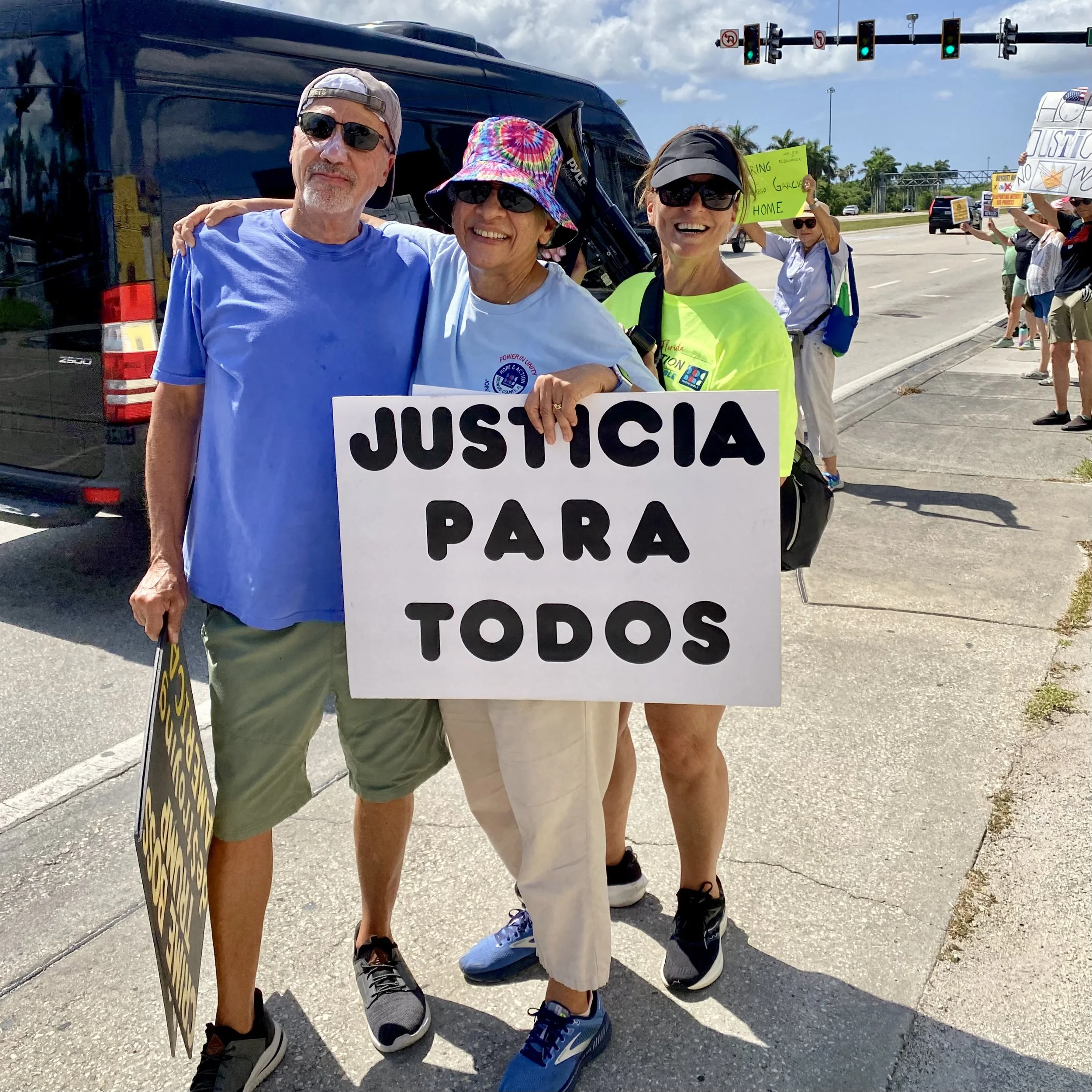 Group of people protesting for justice outside a highway. Three individuals in the front are smiling, wearing sunglasses, and holding a sign that says 'JUSTICIA PARA TODOS'. Several other protesters are in the background with signs, some of which call for justice and others with various messages. The scene appears to take place during the daytime with a cloudy sky.