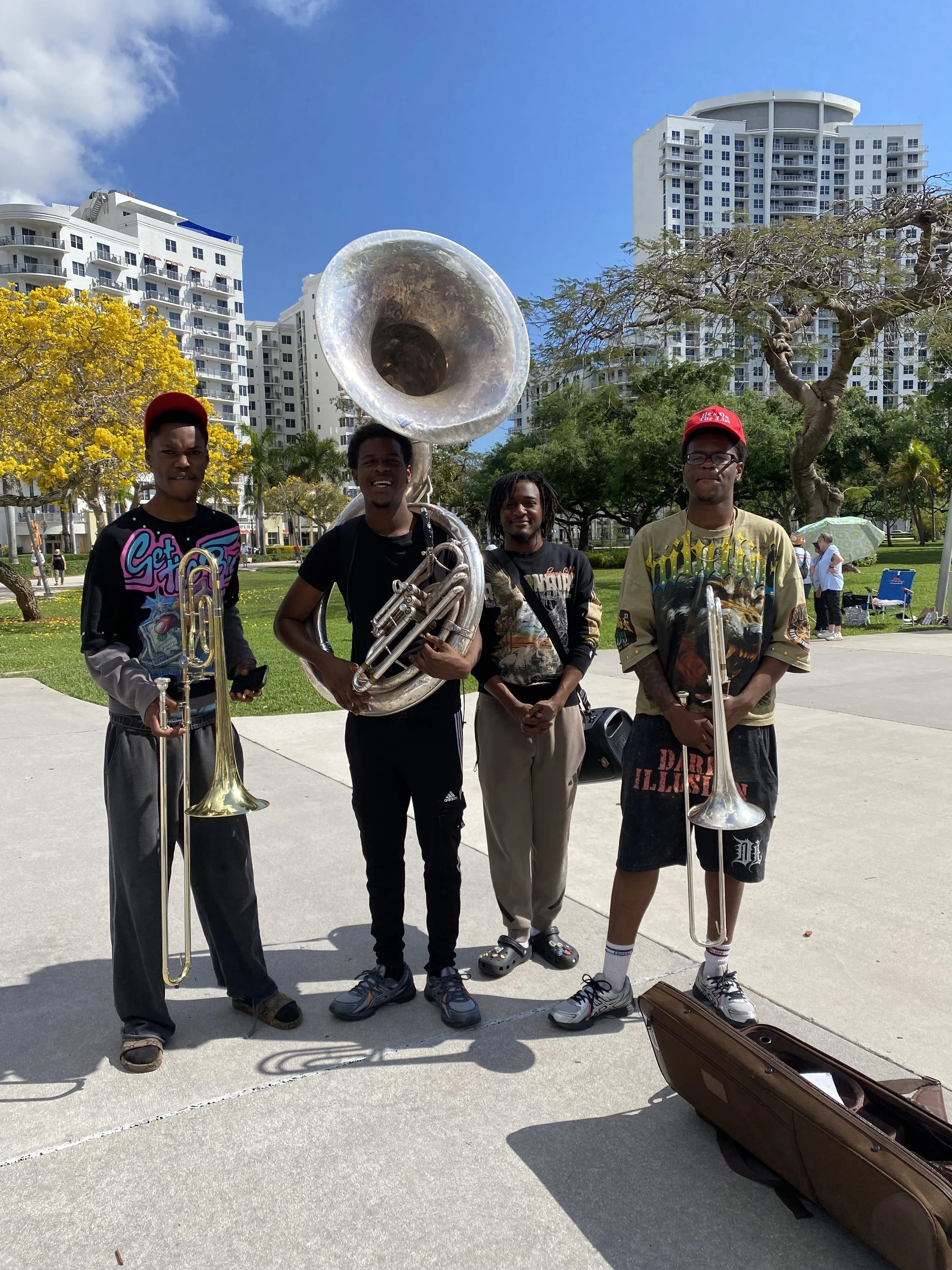 Four men standing outdoors, holding brass musical instruments, with a cityscape and green trees in the background.