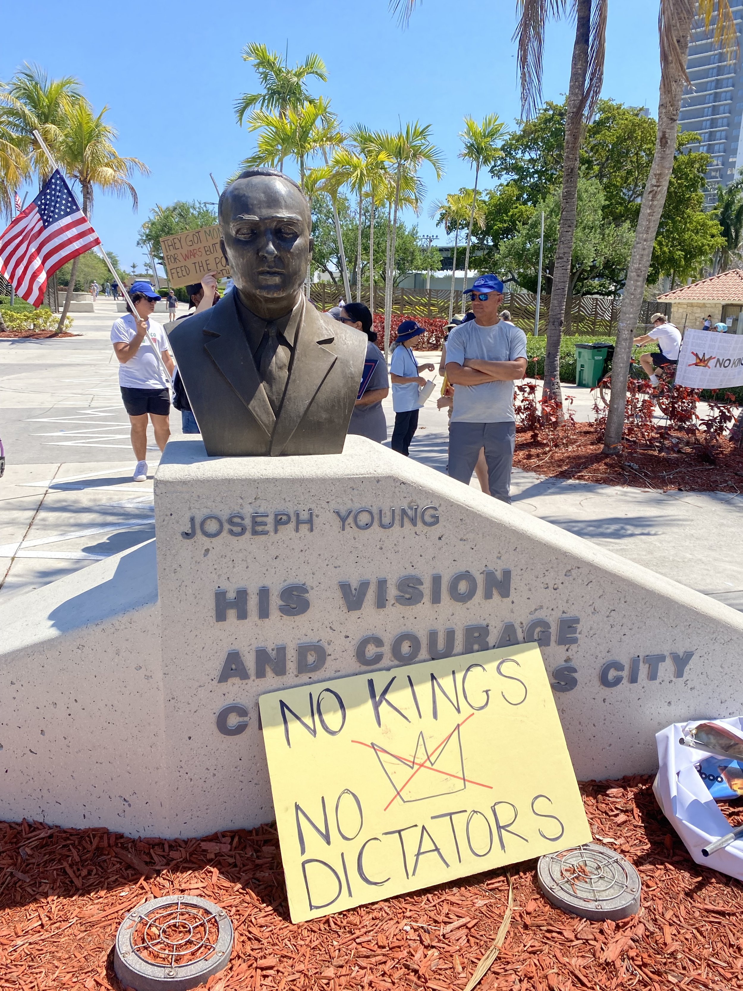 A bust of Joseph Young with a sign that reads, 'HIS VISION AND COURAGE.' In the background, there are people holding signs, one of which says, 'They got money for wars but not for people.' There are American flags, palm trees, and a clear blue sky.