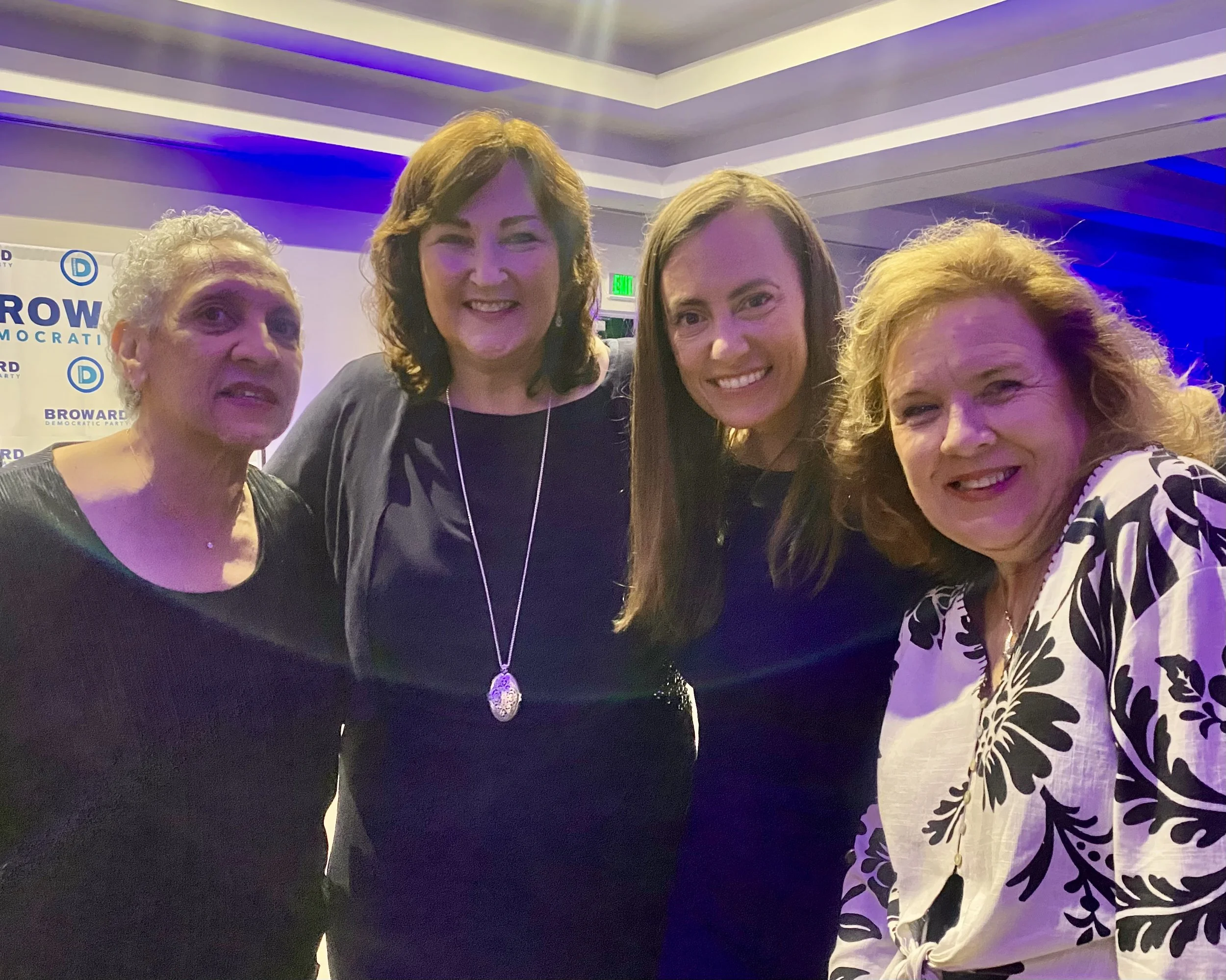 Four women standing closely together at an indoor event with a backdrop that says 'BROWARD DEMOCRATIC PARTY'. They are smiling and posing for the photo, with colorful ceiling lighting in the background.