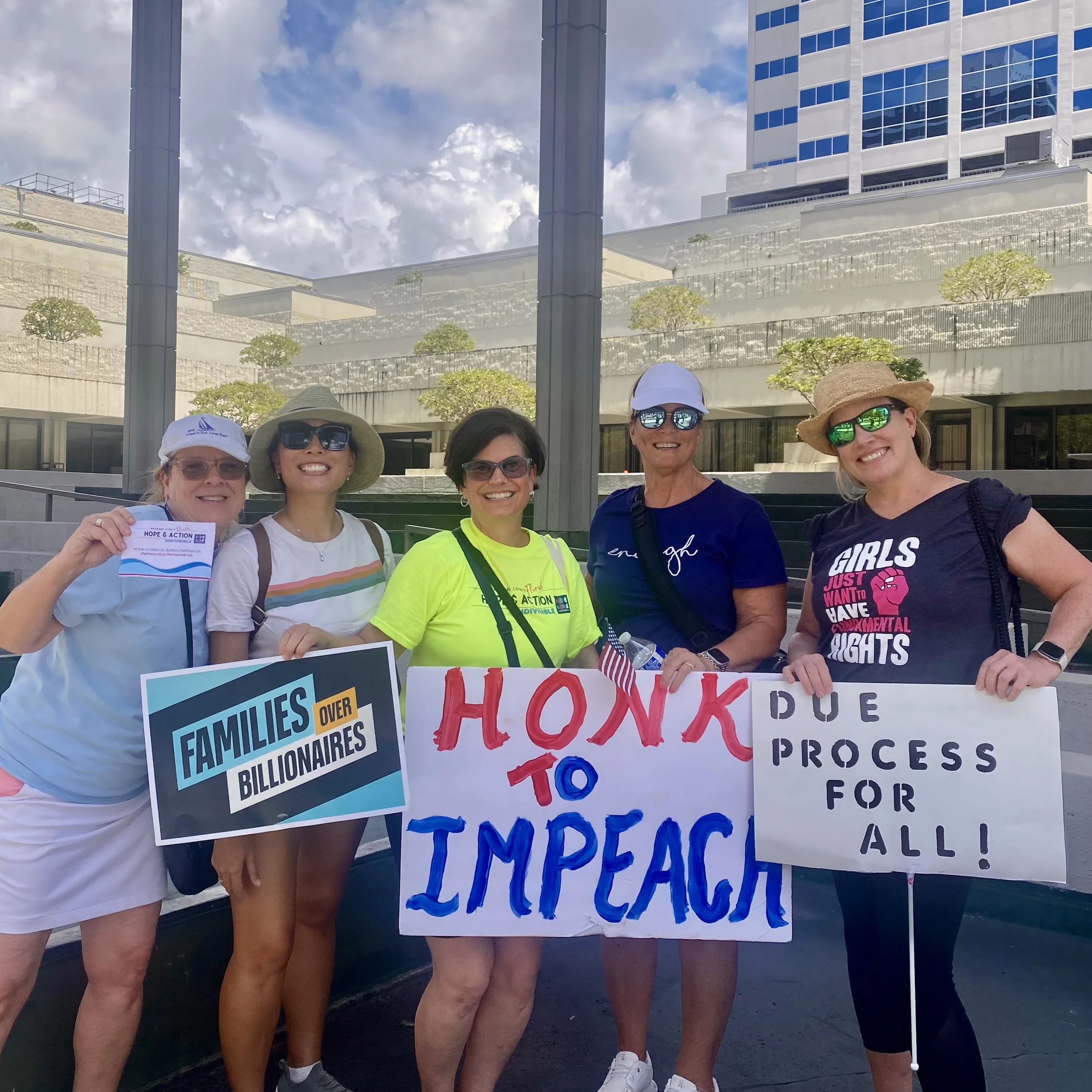 Group of people holding protest signs at an outdoor rally, wearing casual clothes and hats, with buildings and a cloudy sky in the background.