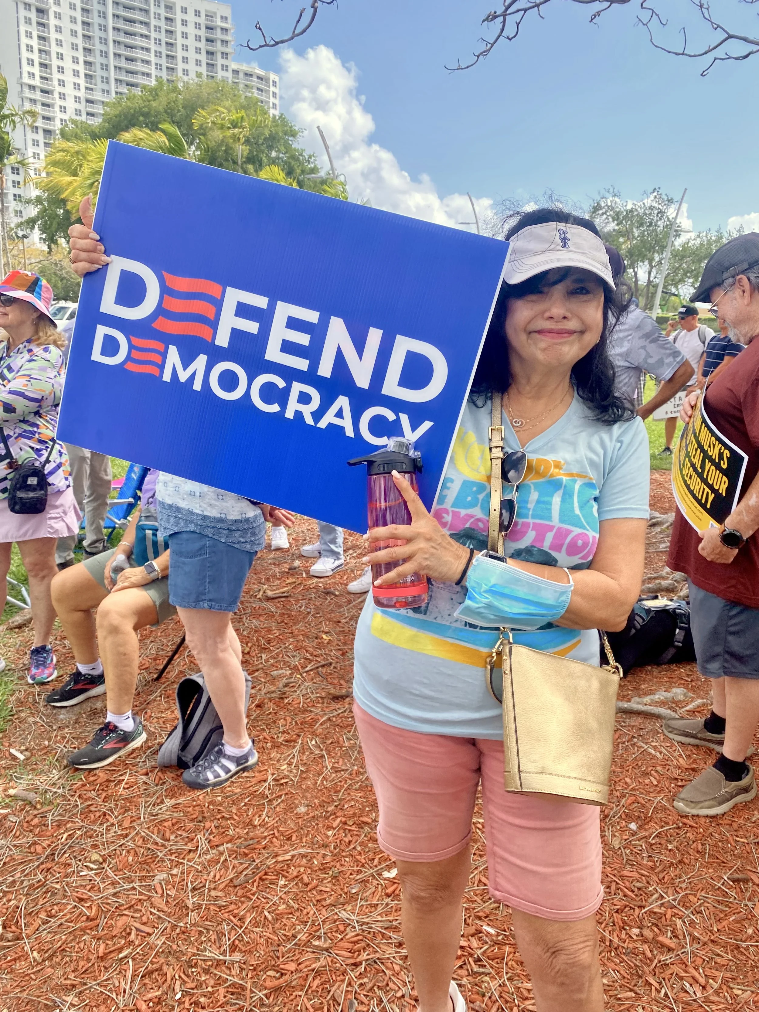 A woman smiling and holding a blue sign that says 'DEFEND DEMOCRACY' at an outdoor event, with other people and tall buildings in the background.