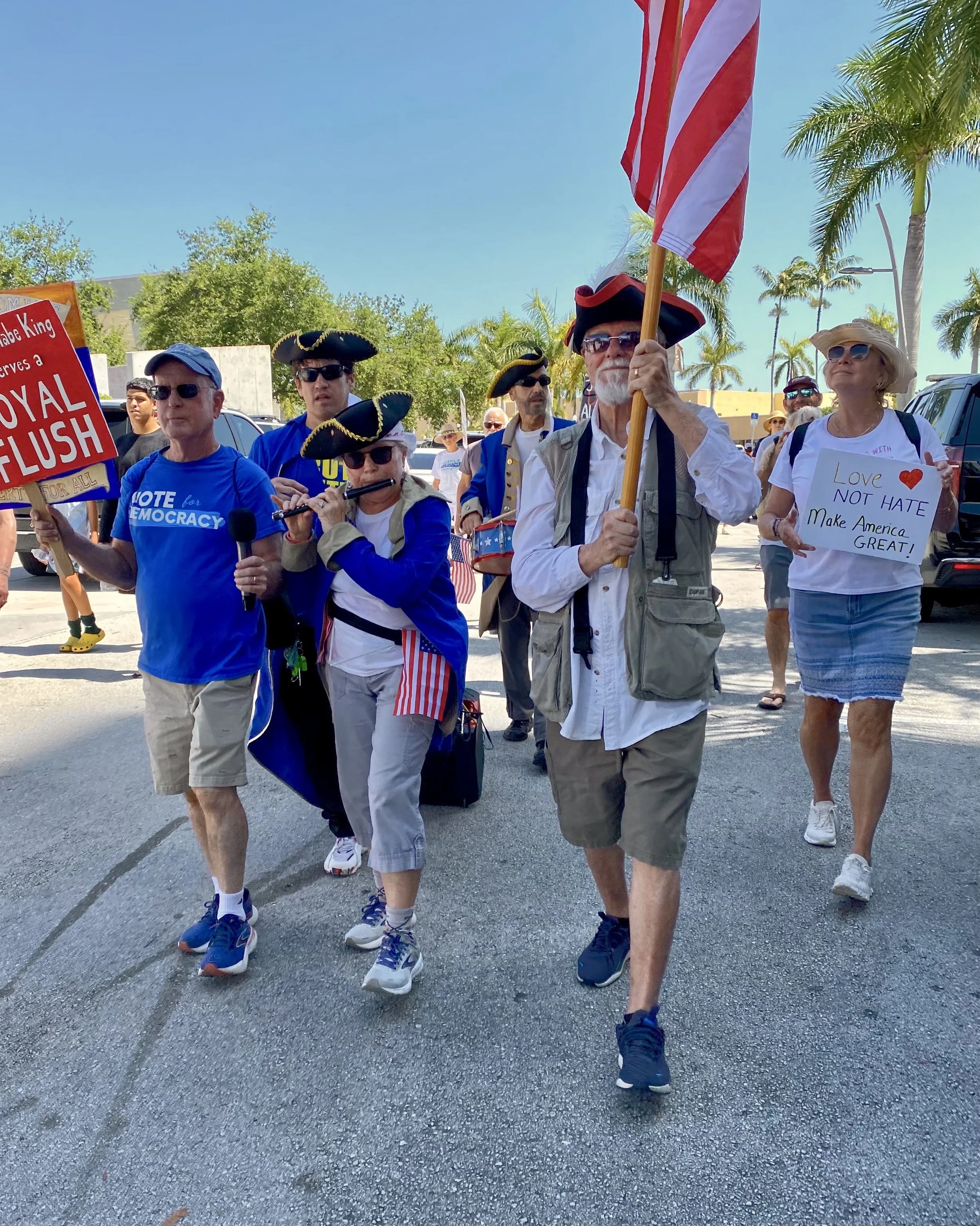 A group of people participating in a parade or march, some wearing colonial-style costumes and tricorn hats, holding signs and flags, with palm trees and a clear blue sky in the background.