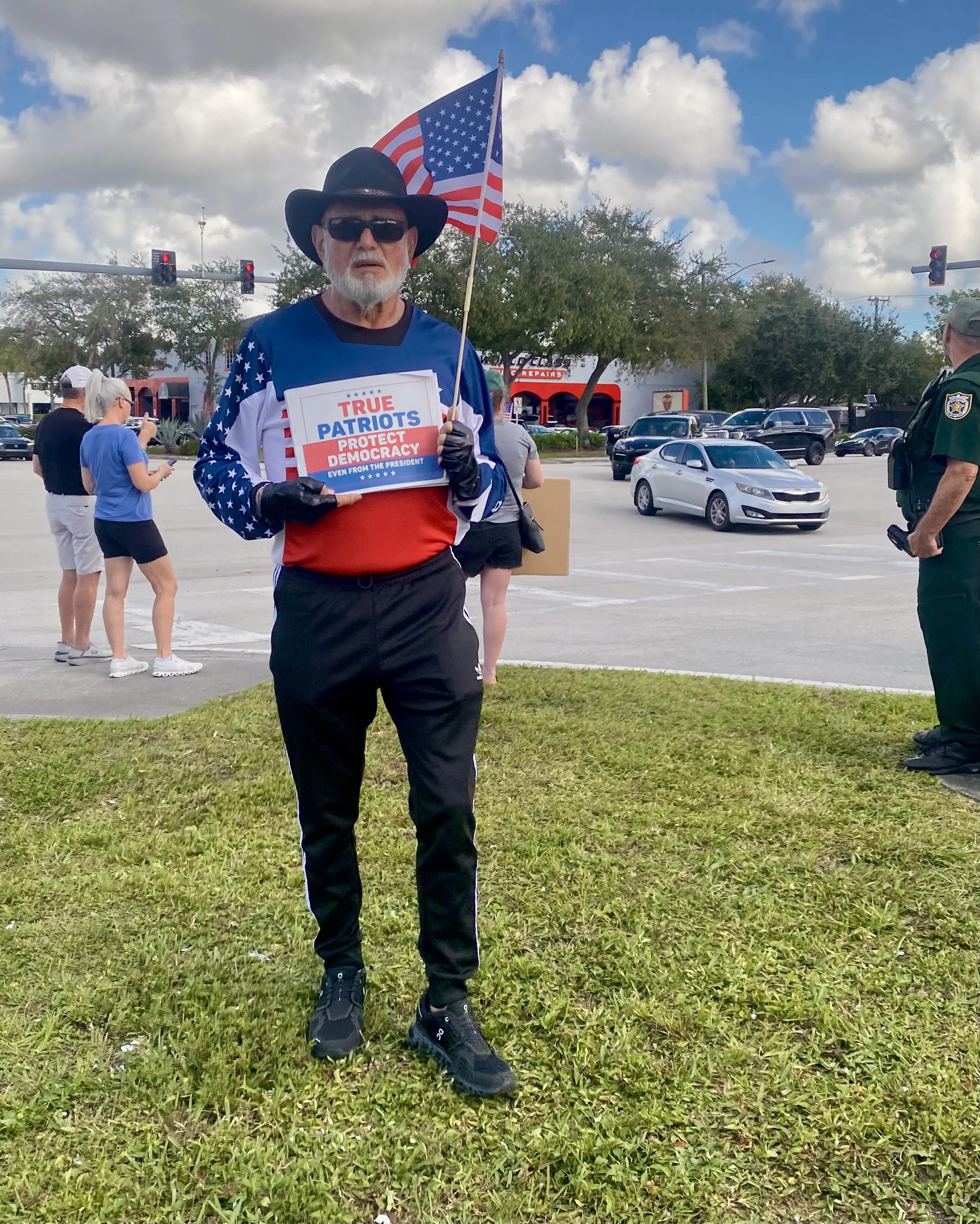 An older man with a beard wearing a black hat, sunglasses, a patriotic jacket, black gloves, and black sneakers holding an American flag and a protest sign that reads "True Patriots Protect Democracy Even from the President" at an outdoor event with a parking lot and cloudy sky in the background.
