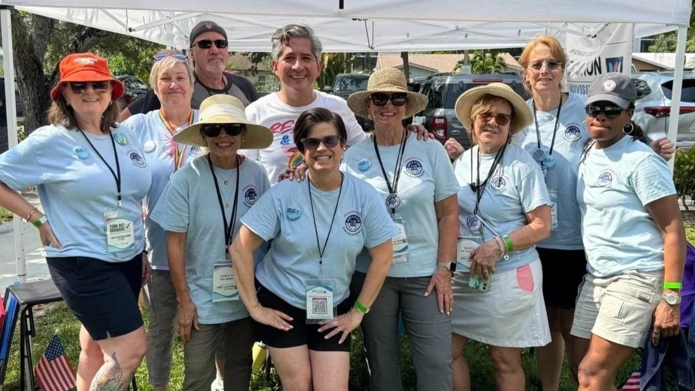 Group of people standing together outdoors under a white tent, some wearing hats and sunglasses, participating in an event or gathering.