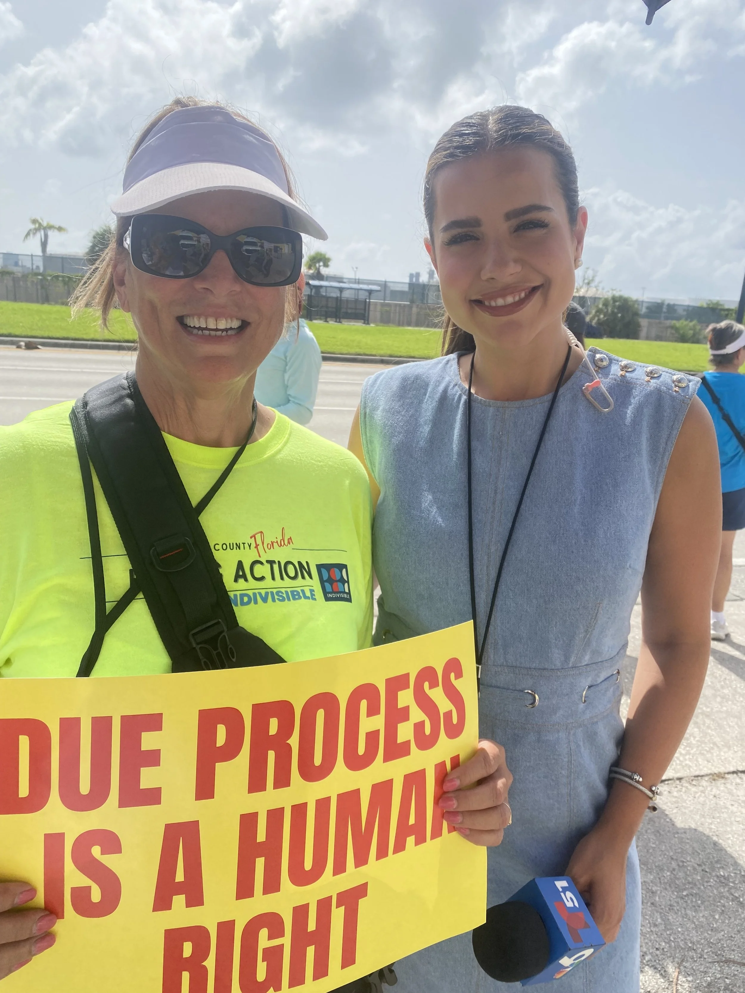 Two women at a protest, one holding a yellow sign with red text, and the other with a gray dress and a microphone. The woman holding the sign wears sunglasses, a baseball cap, and a neon yellow shirt that says 'Florida Action' and 'Indivisible.' The woman in the dress is smiling, with her hair tied back, and has a microphone labeled '5' near her. Background includes a sidewalk, a grassy area, and two other people.