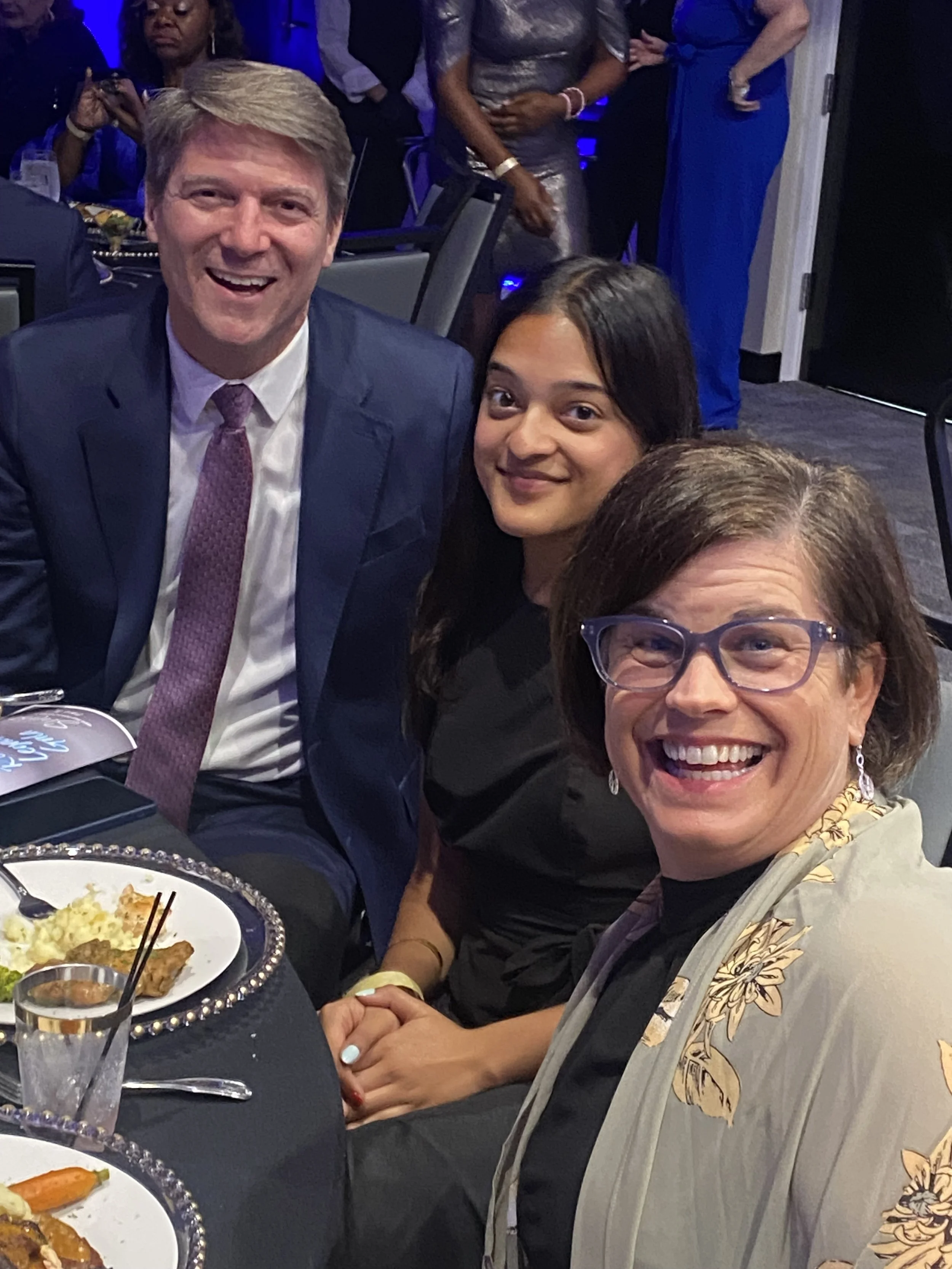People smiling and holding hands at a dinner event, with plates of food on the table and a woman wearing glasses at the front.