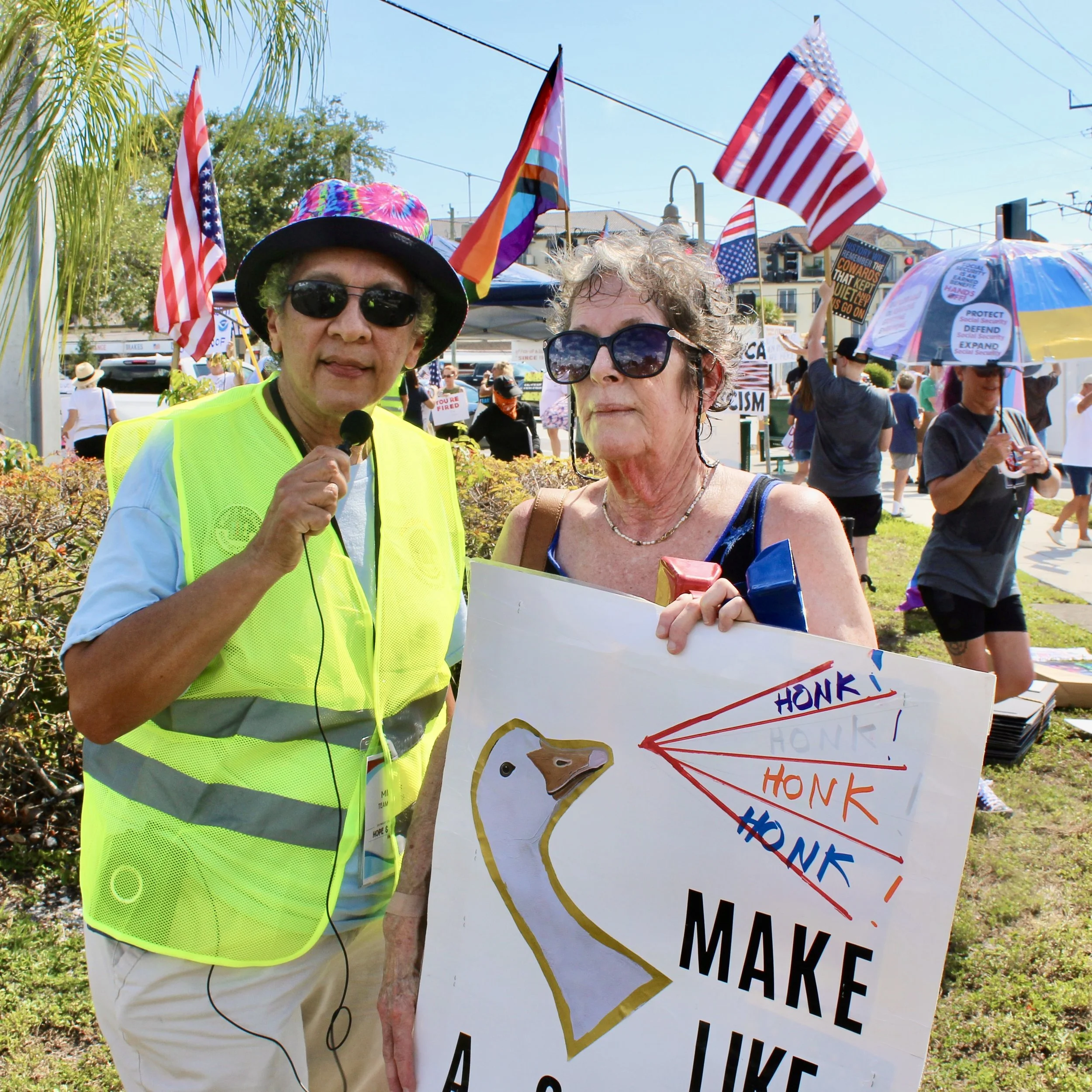 Two women are participating in a protest or rally outdoors on a sunny day. One woman, wearing sunglasses, a bright yellow vest, and a tie-dye hat, holds a microphone. The other woman, also wearing large sunglasses, is holding a protest sign with colorful writing and illustrations. Flags, including American and rainbow LGBTQ pride flags, are visible in the background along with other protesters holding signs and umbrellas.