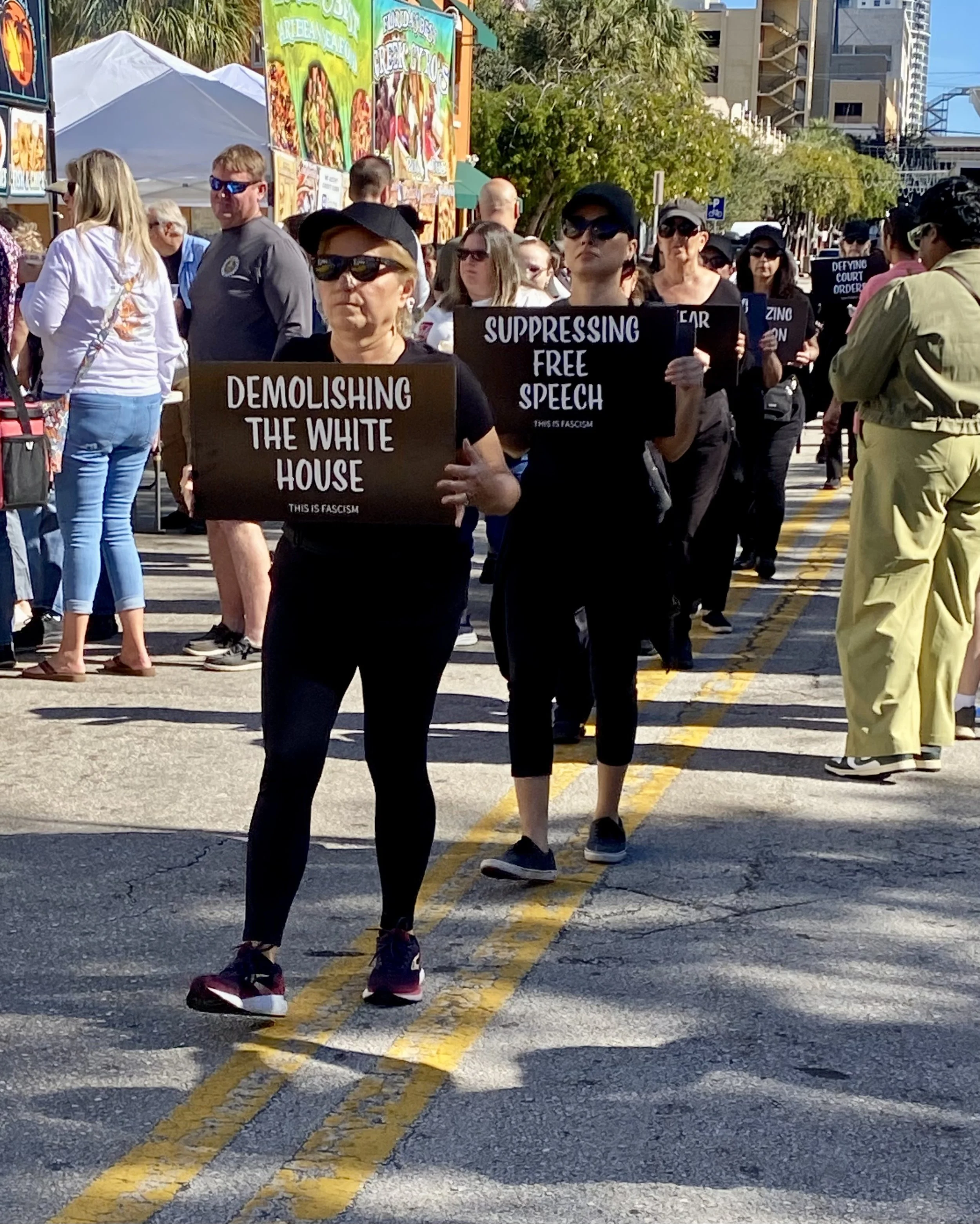 Protestors marching on a city street holding signs with messages about social and political issues, such as ending White House demolishing and supporting free speech, with tall buildings and trees in the background.