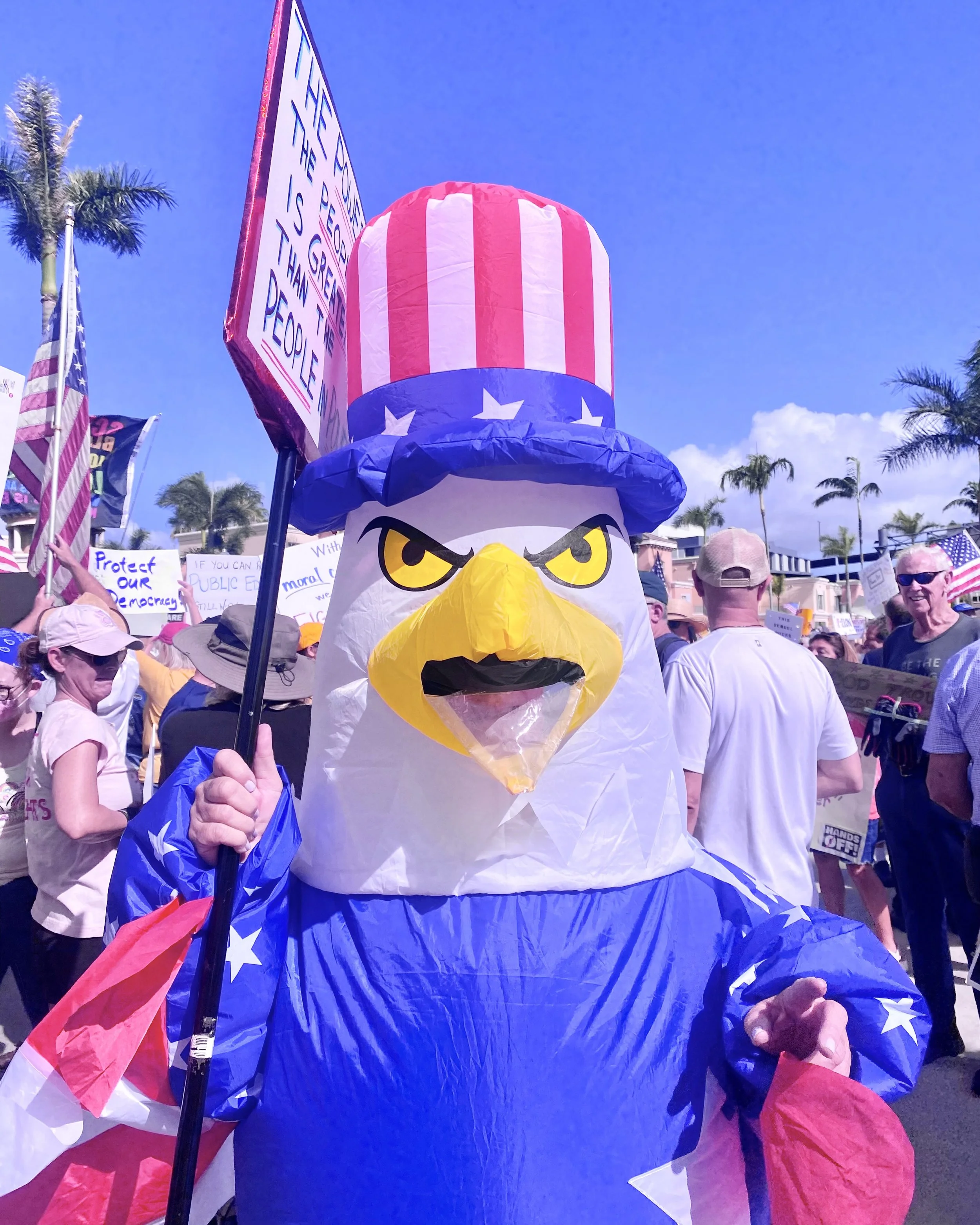 Person wearing a Bald Eagle costume with an Uncle Sam hat at a parade or rally, holding a flag with stars and stripes, with a crowd and signs in the background.