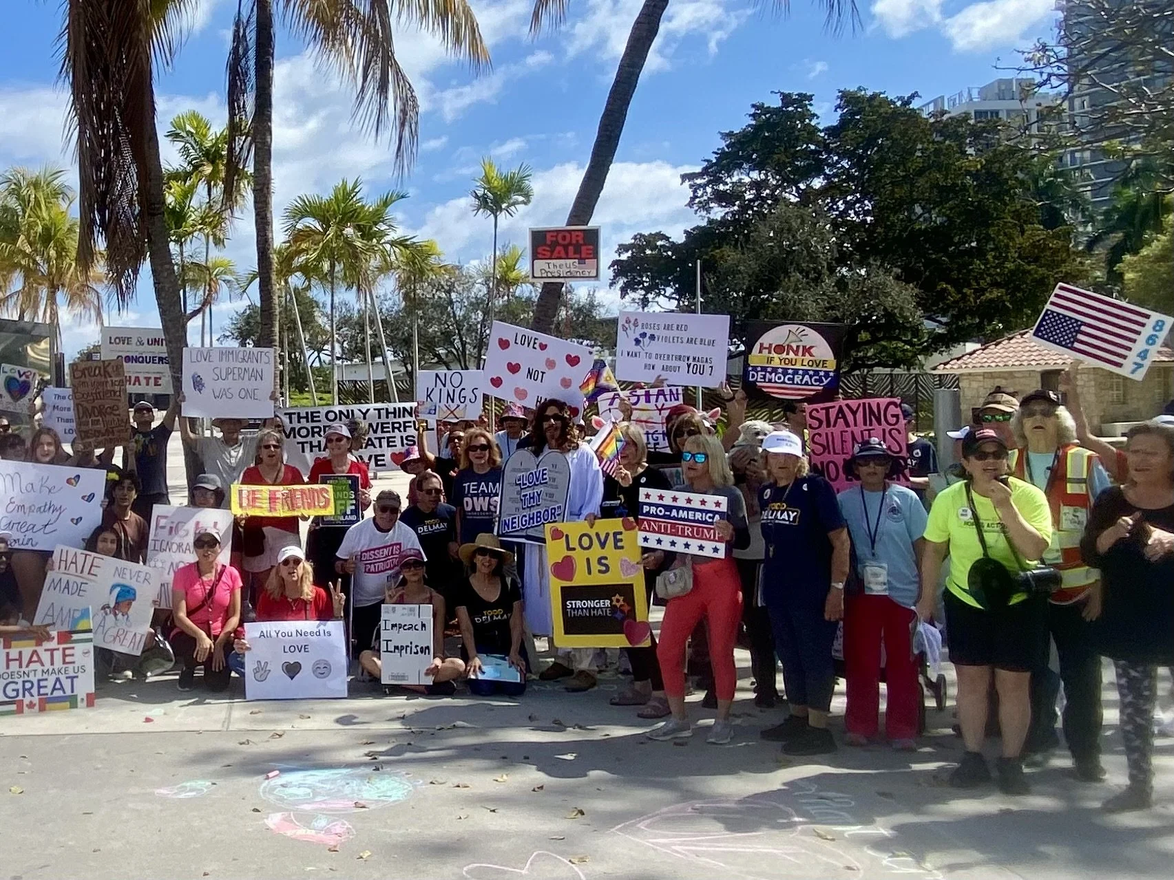 A diverse group of people participating in a protest or march, holding various signs and banners advocating for love, equality, and social justice. The crowd includes men and women, some wearing sunglasses, hats, and colorful clothing, standing outside on a sunny day with palm trees and a building in the background.
