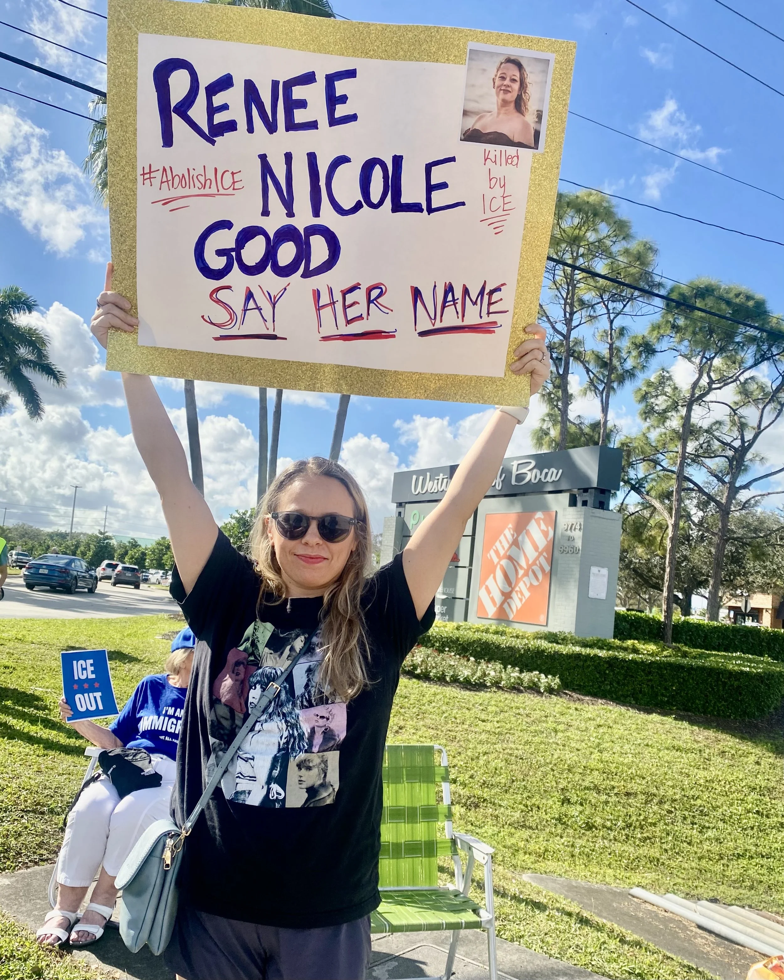 A woman at a protest holding a sign with a photo of Renee Nicole, with the text 'Renee Nicole Good'. The sign includes handwritten notes saying '#Abolish ICE' and 'killed by ICE'. Another person in blue is sitting in the background holding a sign that says 'ICE OUT'. The scene is outdoors with trees, a blue sky, and a car lot in the background.