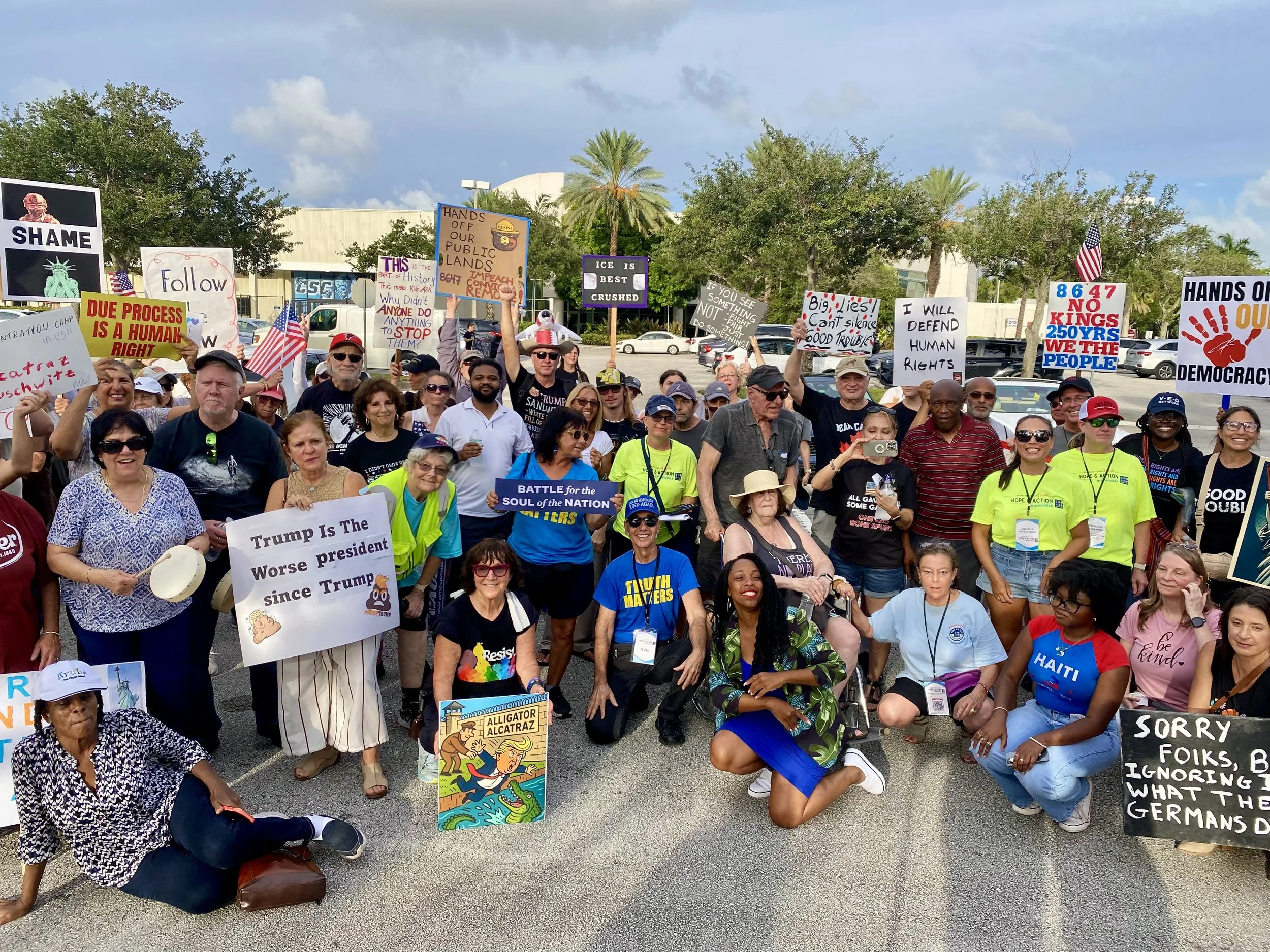 A diverse group of people gathered outdoors at a protest or rally, holding various signs with messages about politics, democracy, and social issues. Some signs include slogans like 'DUE PROCESS IS A HUMAN RIGHT,' 'IF YOU SEE SOMETHING NOT FIGHT FUTURE SOMETHING,' and 'HANDS OFF OUR LAND.' There are American flags, clear skies, trees, and a parking lot in the background.