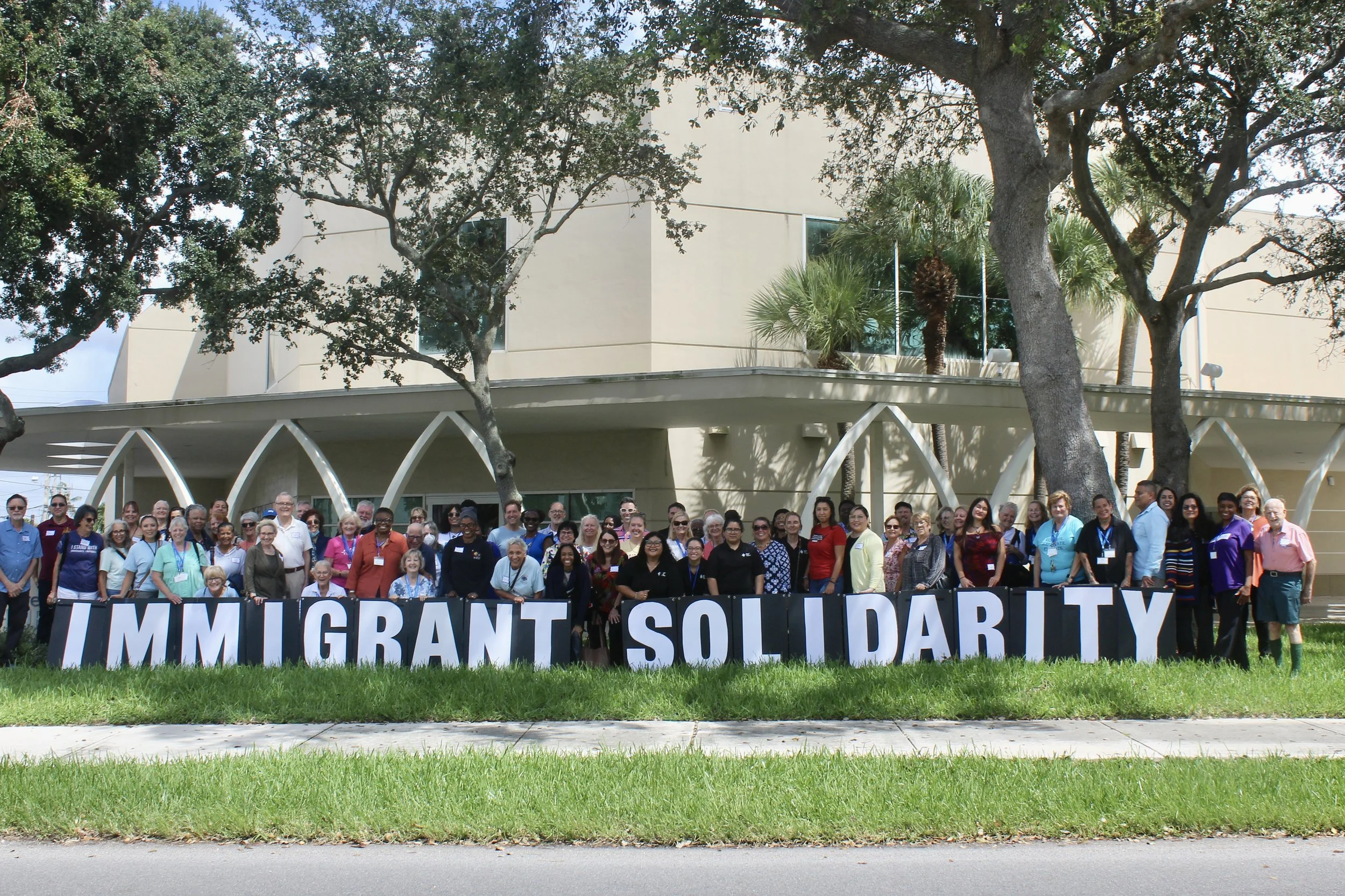 Large group of people standing outdoors behind a banner that reads 'IMMIGRANT SOLIDARITY'. The group is in front of a modern building with large windows and trees. The sky is partly cloudy.