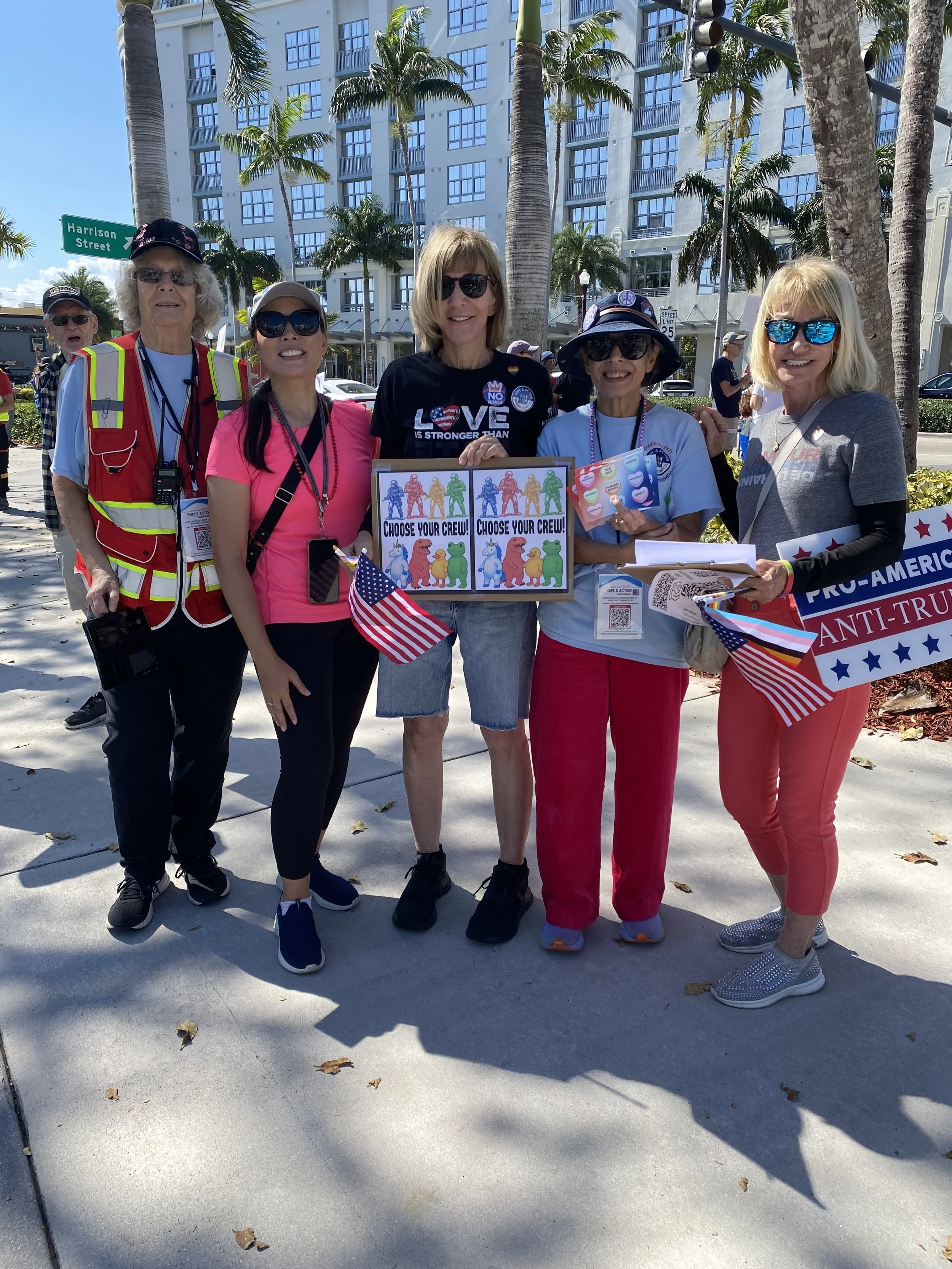 A group of five women standing outdoors on a sunny day, holding signs and American flags, with tall palm trees and a large building in the background, appearing at a rally or protest.