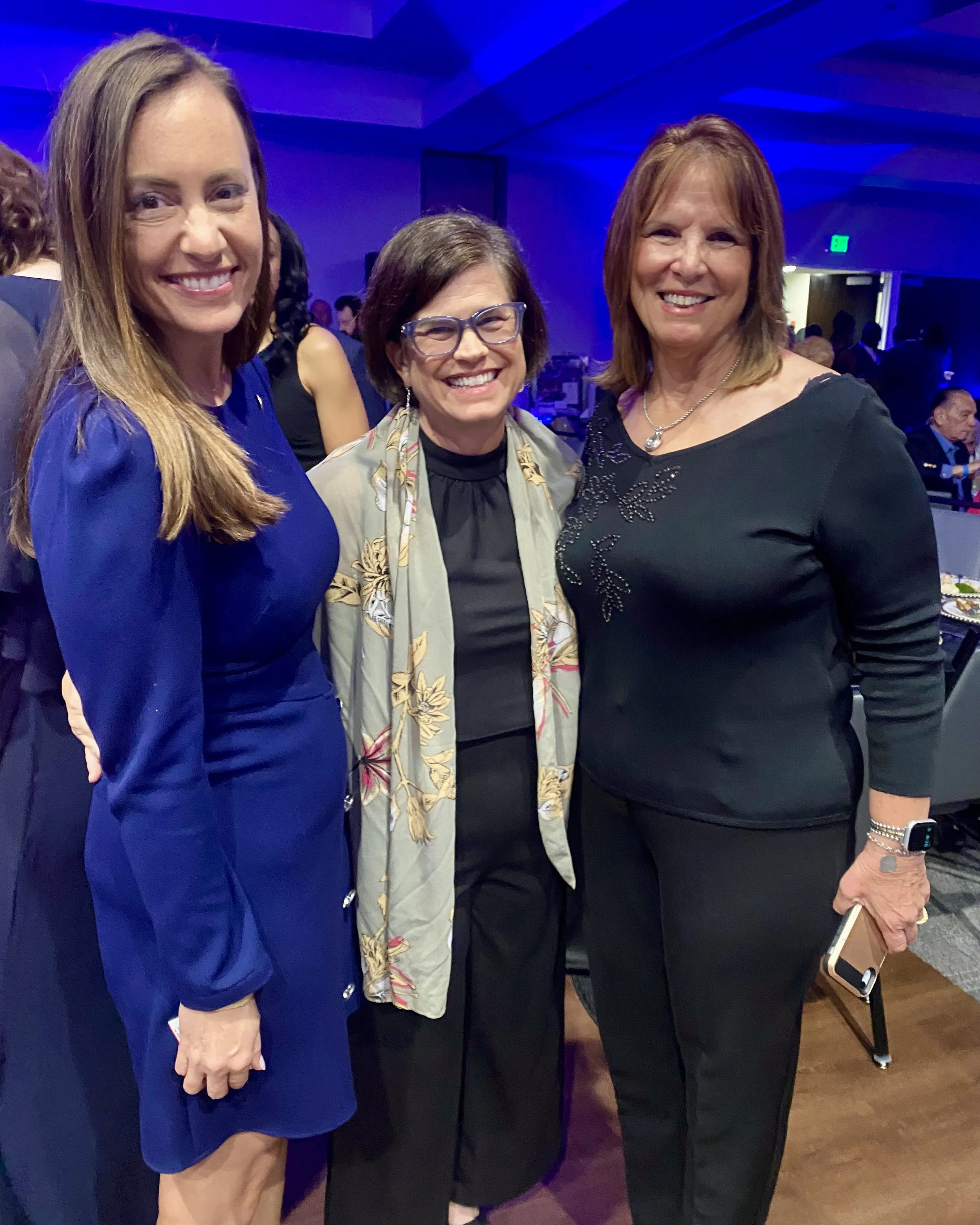 Three women posing at an indoor event with blue lighting. The woman on the left has long brown hair and is wearing a blue dress. The woman in the middle has short dark hair, glasses, and a floral shawl. The woman on the right has shoulder-length reddish hair and is wearing a black dress with a necklace. They are smiling and standing close together.