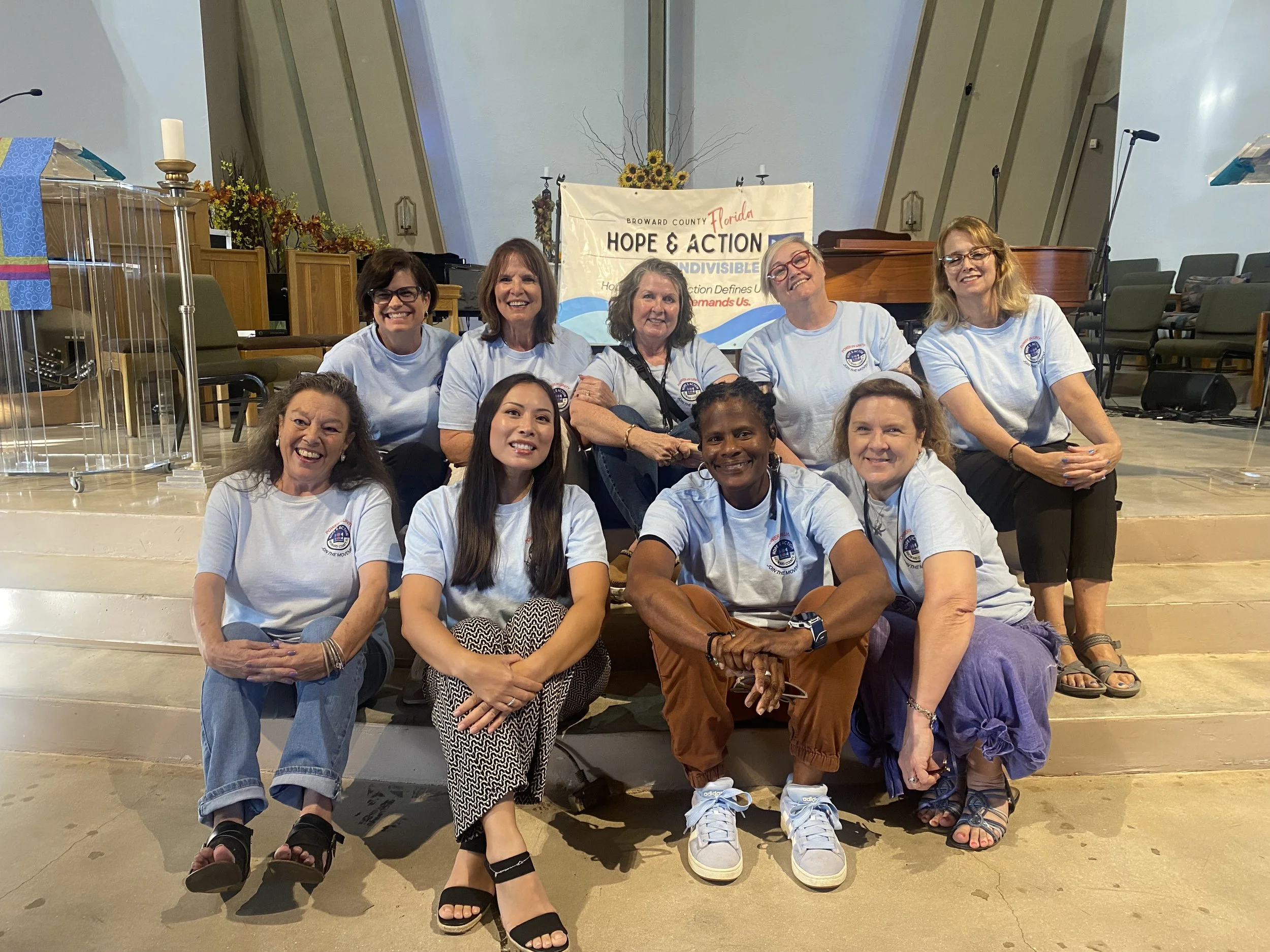 A group of women sitting and kneeling in front of a banner that reads 'Hope & Action' at a church or community center. The women are smiling and wearing matching light blue T-shirts, with some wearing glasses. The background includes chairs, a piano, and some floral arrangements.