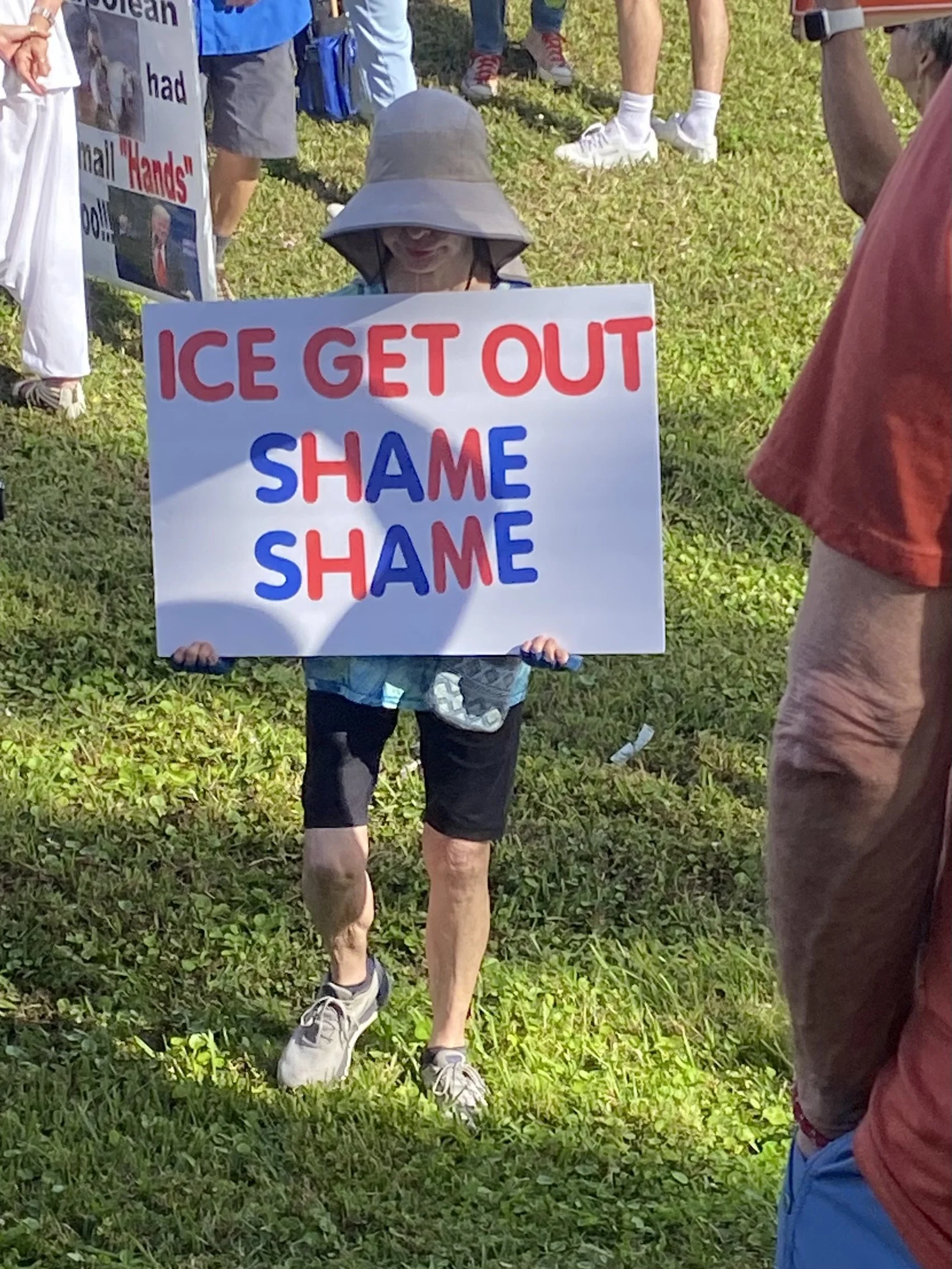 A person is holding a sign protesting climate change, with the words 'ICE GET OUT SHAME SHAME' in red and blue letters. The person is wearing a wide-brimmed hat, shorts, and sneakers, and is standing on grass. Other people are visible in the background, some wearing athletic clothing.