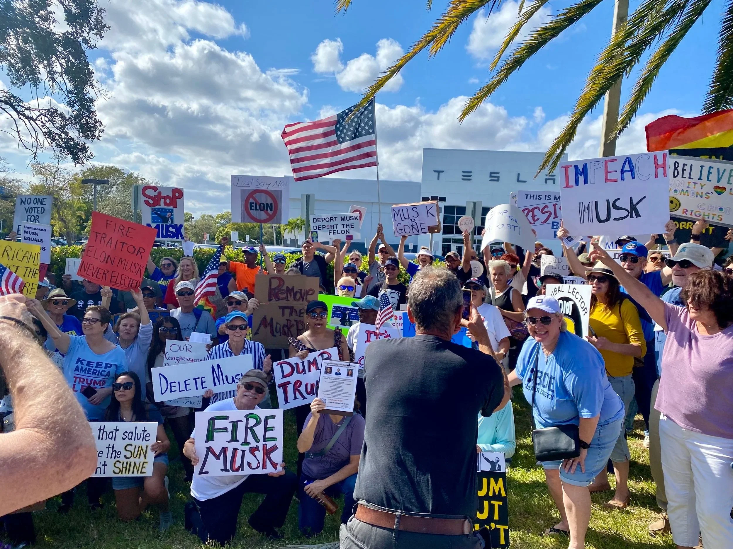 A crowd of people holding various protest signs outside in front of a Tesla building, with palm trees and cloudy sky in the background.