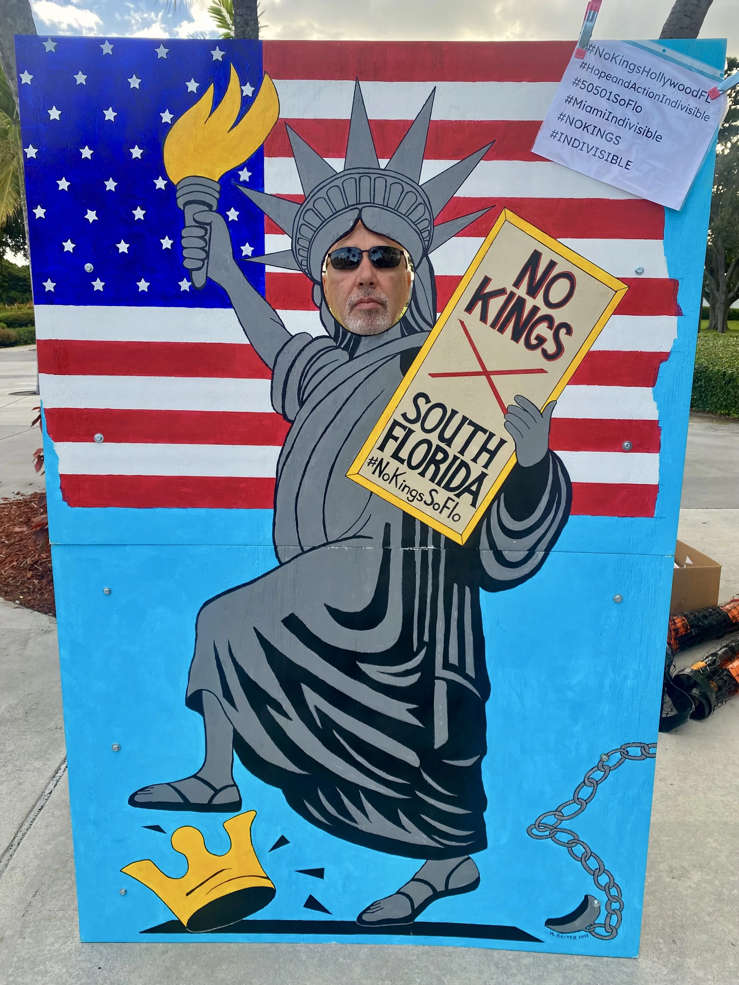 A painted cutout of the Statue of Liberty holding a 'No Kings' sign, with a background resembling the American flag, and a person’s face visible through the statue's face opening.