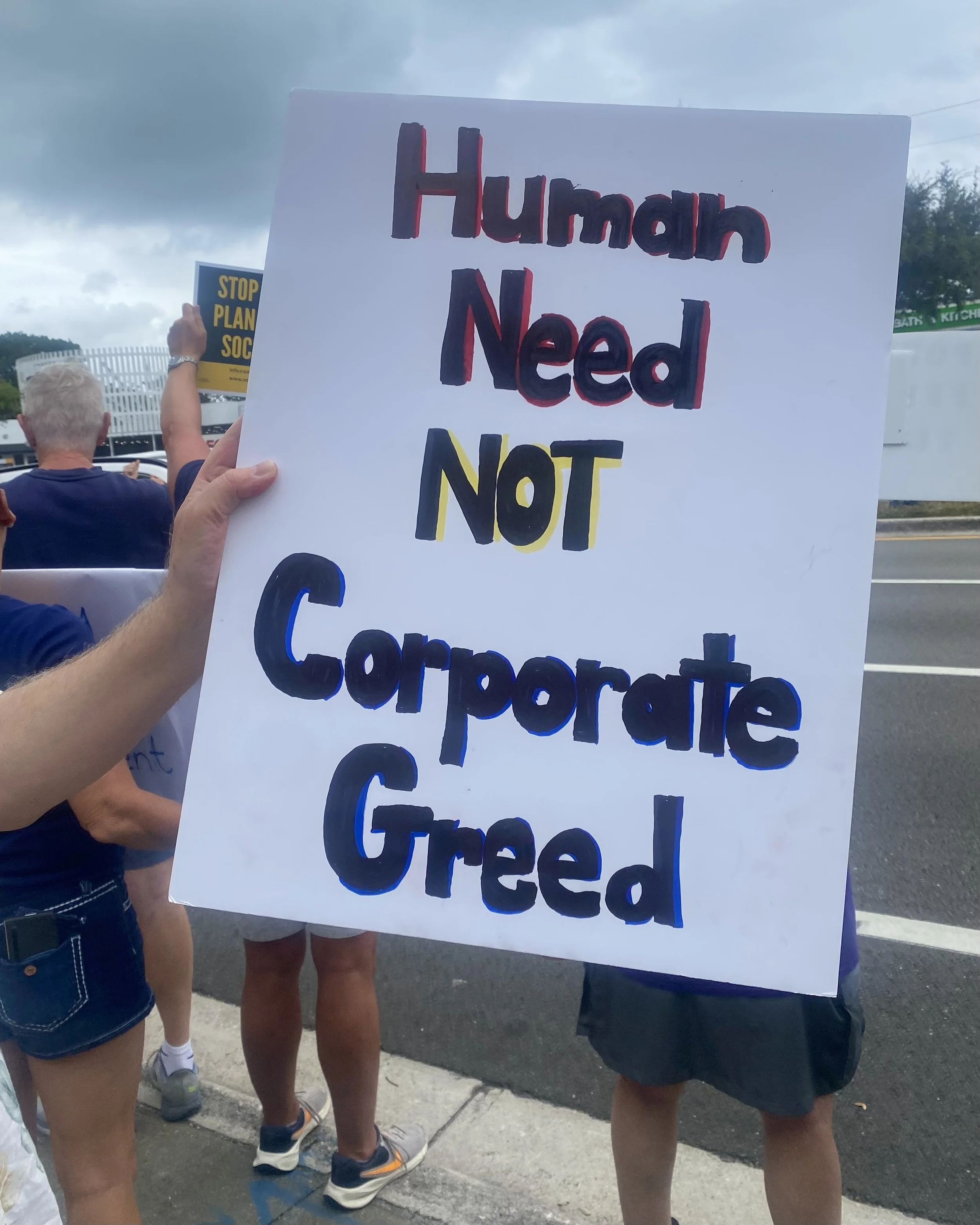 Protesters holding signs at a public demonstration; one sign reads "Human Need NOT Cooperation" with the word "NOT" highlighted in yellow