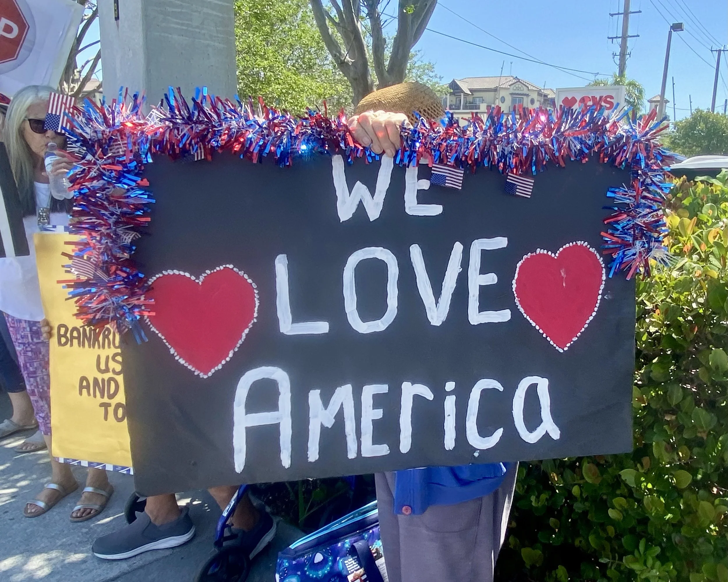 A person holding a sign with writing that says "We Love" and appears to be part of a larger message. The sign is decorated with red, white, and blue tinsel and small American flags, and appears to be at an outdoor event with other people and buildings in the background.