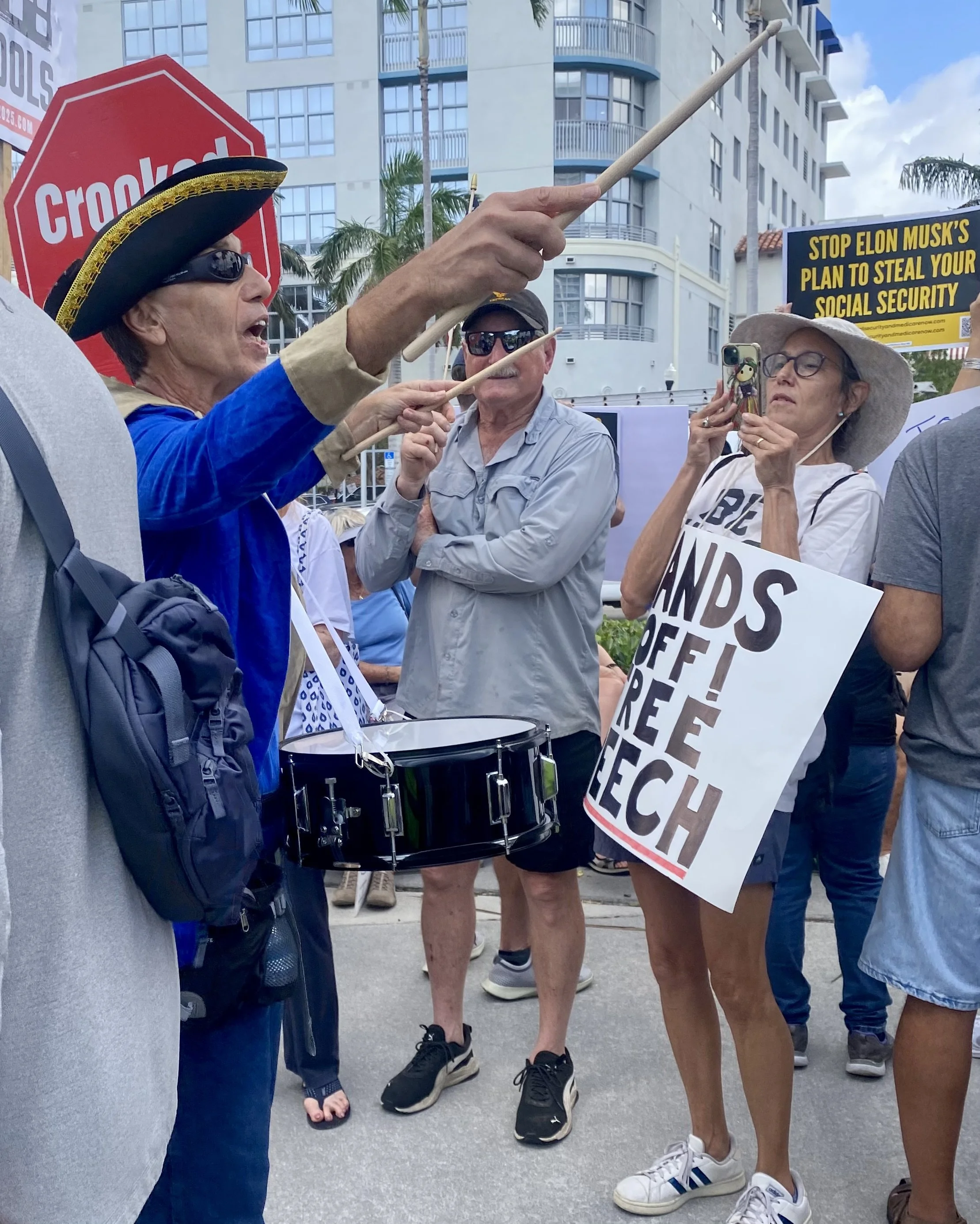 Group of protesters at an outdoor rally, with one woman wearing a pirate hat and sunglasses, speaking passionately and gesturing with drumsticks. Another woman holds a phone, capturing a video, while holding a sign that reads 'Stop Elon Musk's plan to steal your social security.' Several other people are visible in the background, some wearing sunglasses and casual clothing. The setting is an urban area with modern buildings and palm trees.