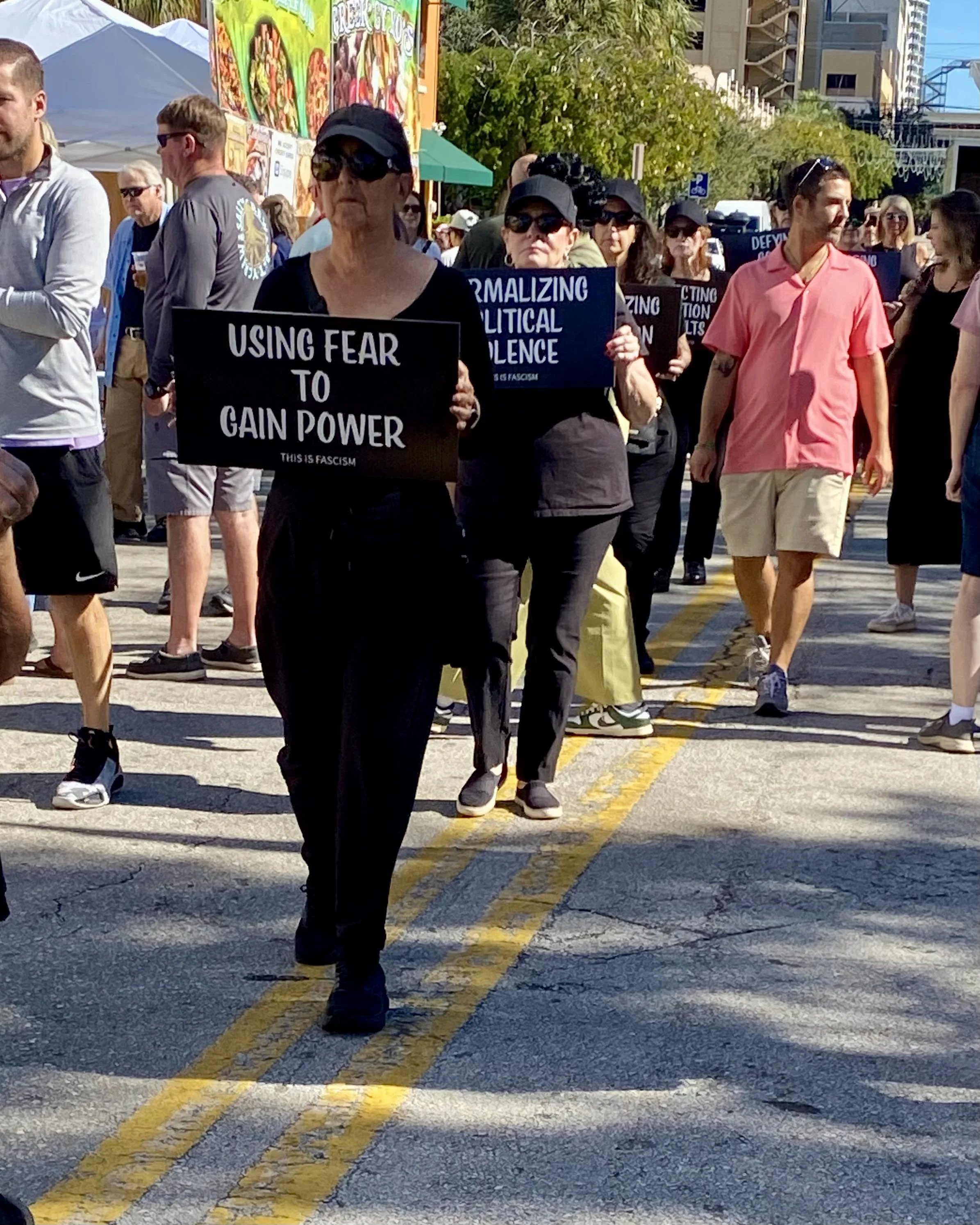 Protesters marching on a city street holding signs with messages about political issues, with some wearing sunglasses and casual clothing. The scene appears to be during daylight with urban buildings and trees in the background.
