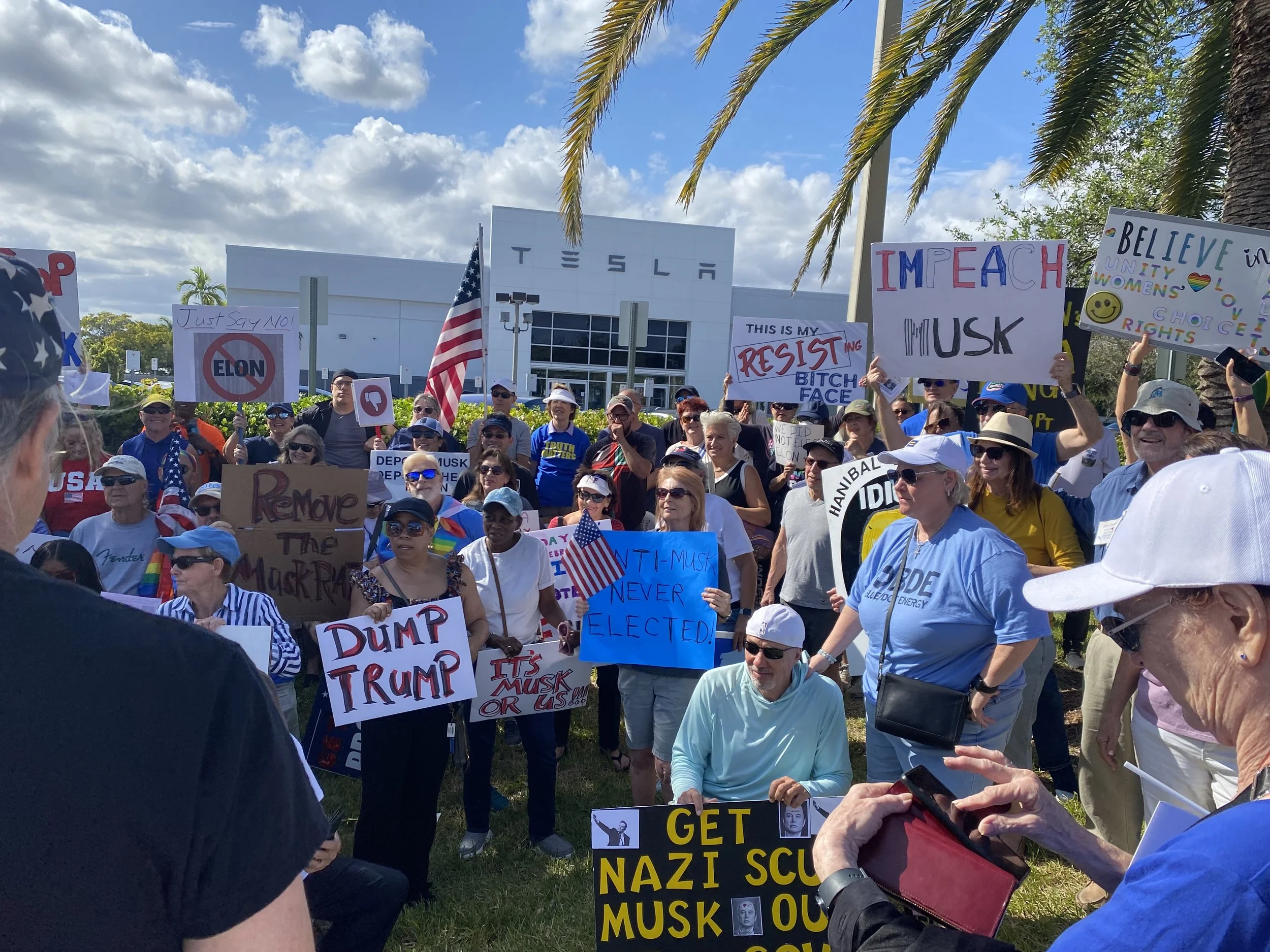Group of people holding protest signs at a rally or demonstration outdoors, with trees and a clear blue sky in the background.