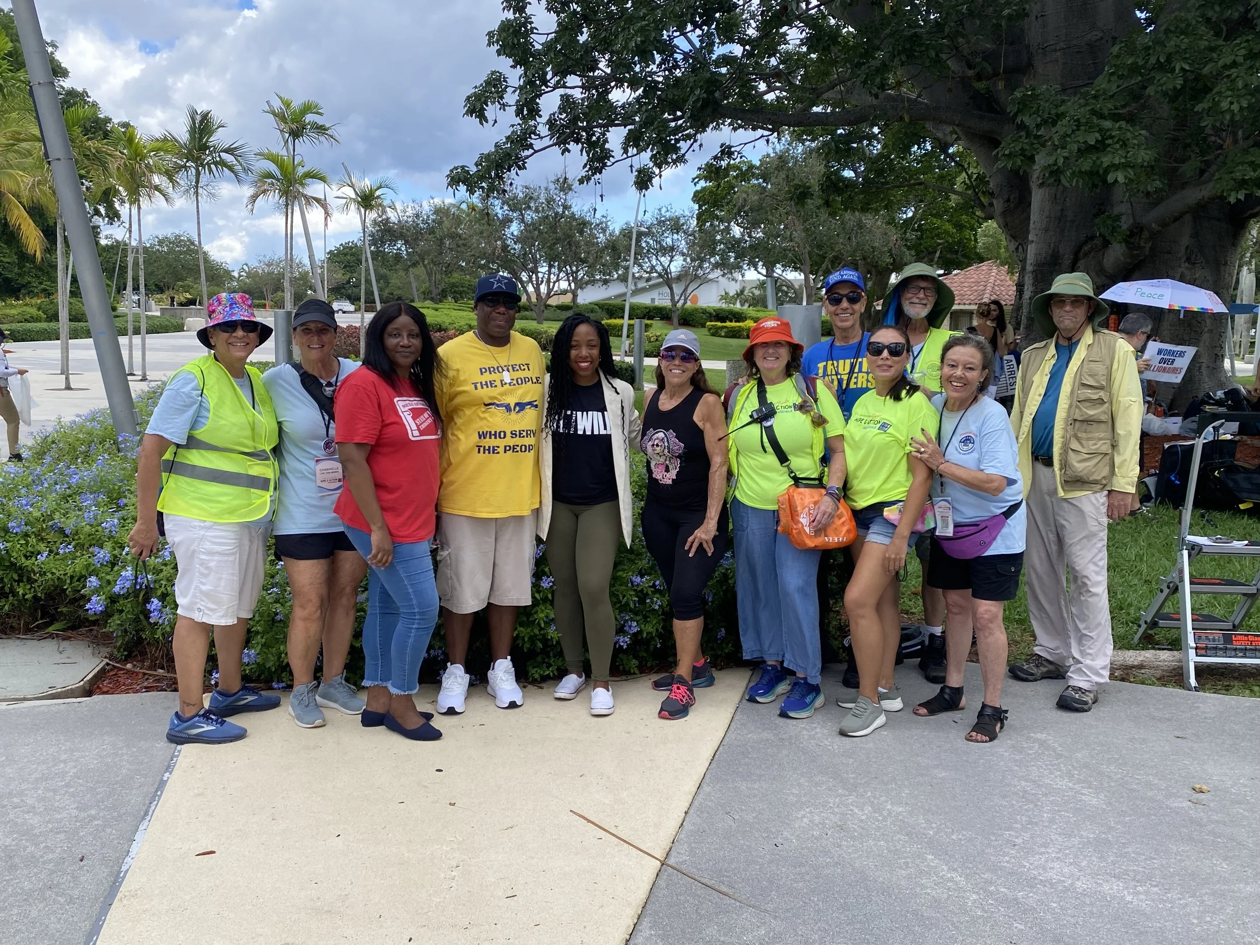 A group of diverse people standing outdoors under a large tree, some wearing brightly colored shirts and hats, with a background of palm trees, shrubs, and a building.