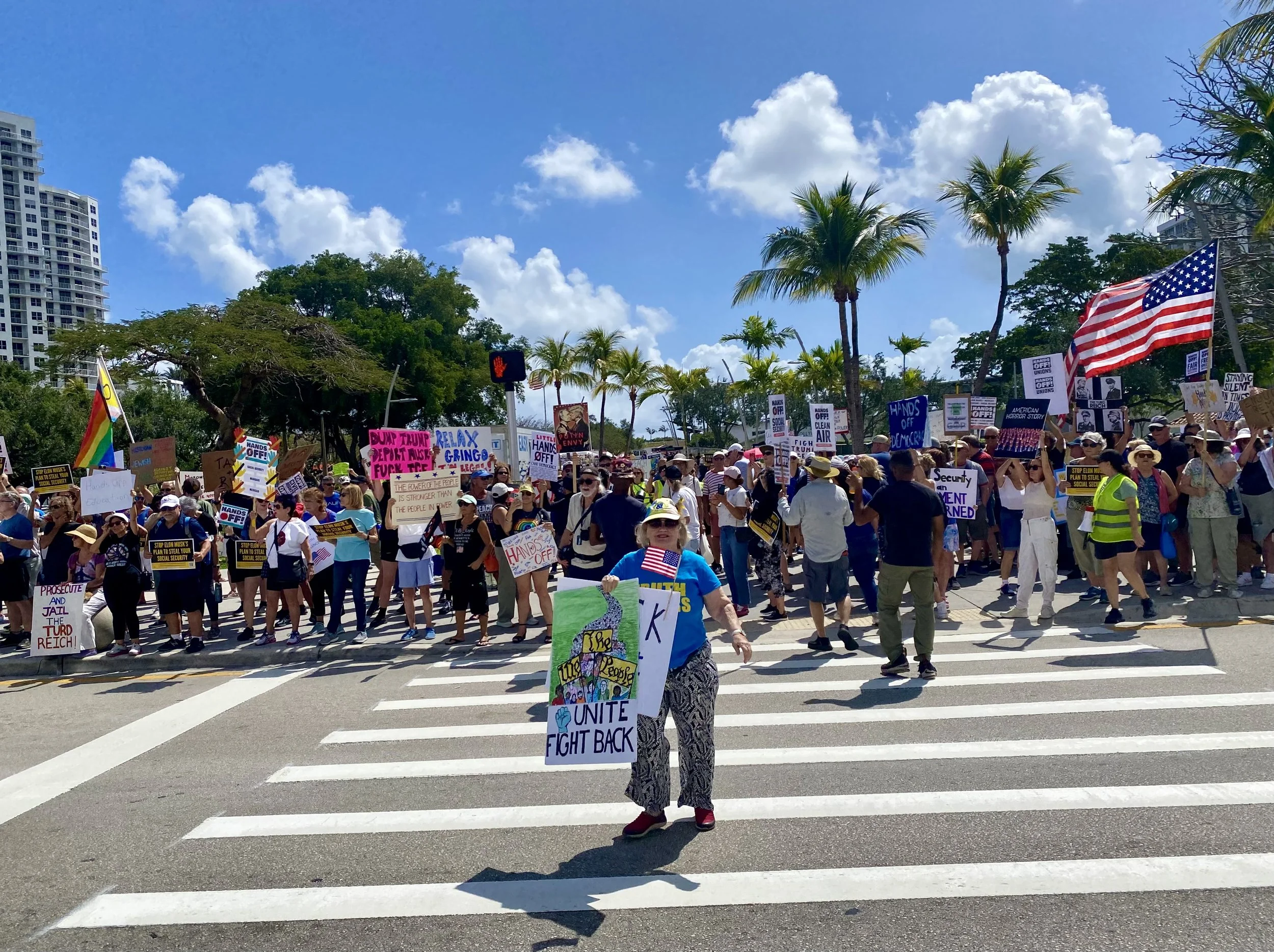 A large group of people protesting outdoors, holding various signs and flags, including a rainbow pride flag and an American flag, under a blue sky with scattered clouds, with trees and city buildings in the background.