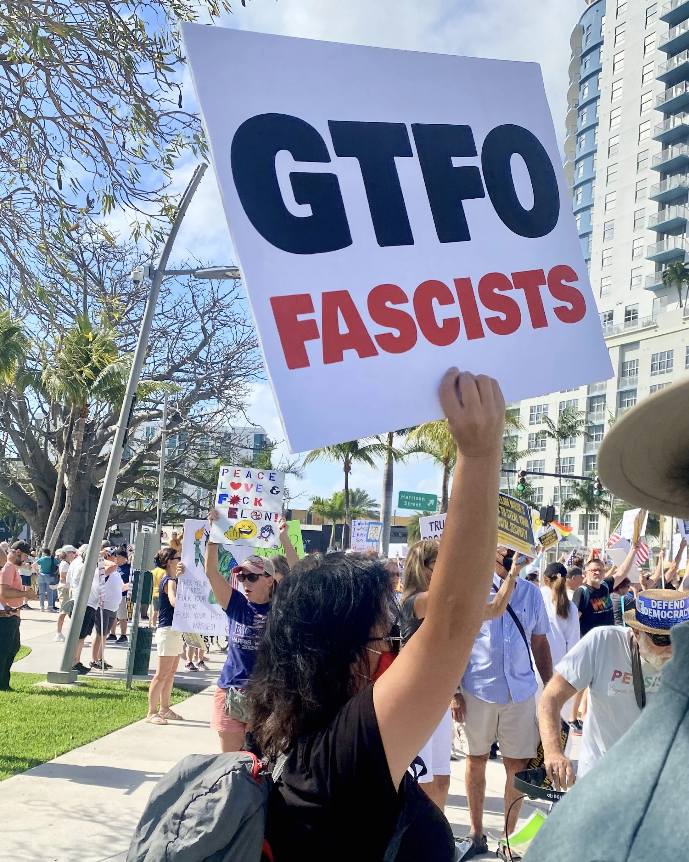Protesters at an outdoor rally holding signs and banners, with a cityscape of high-rise buildings, trees, and a street sign in the background.