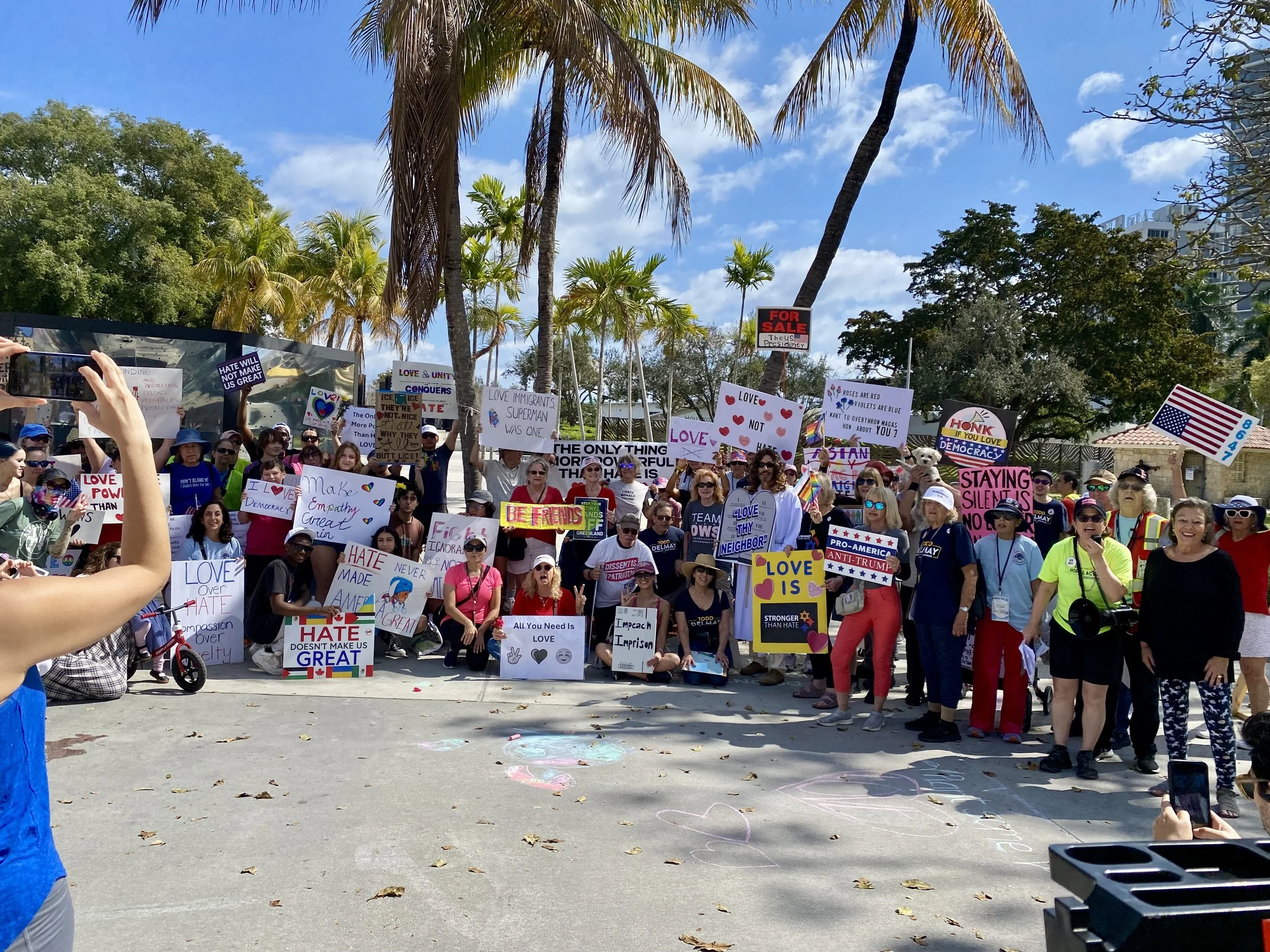 Large crowd gathered outdoors holding various protest signs with messages advocating love, unity, and anti-hate, under palm trees and a partly cloudy sky.