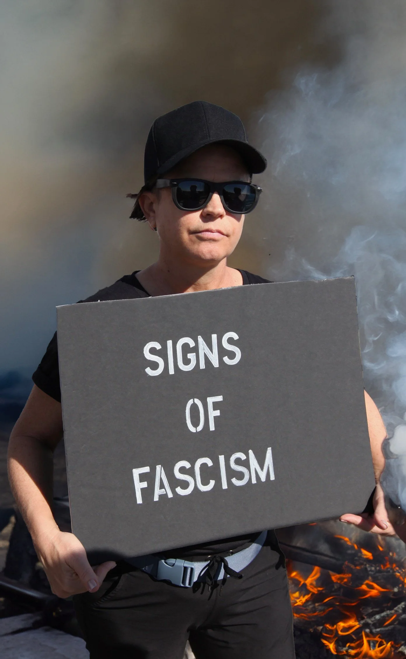 A woman wearing sunglasses and a cap holding a sign that reads 'SIGNS OF FASCISM' while standing in front of a fire.