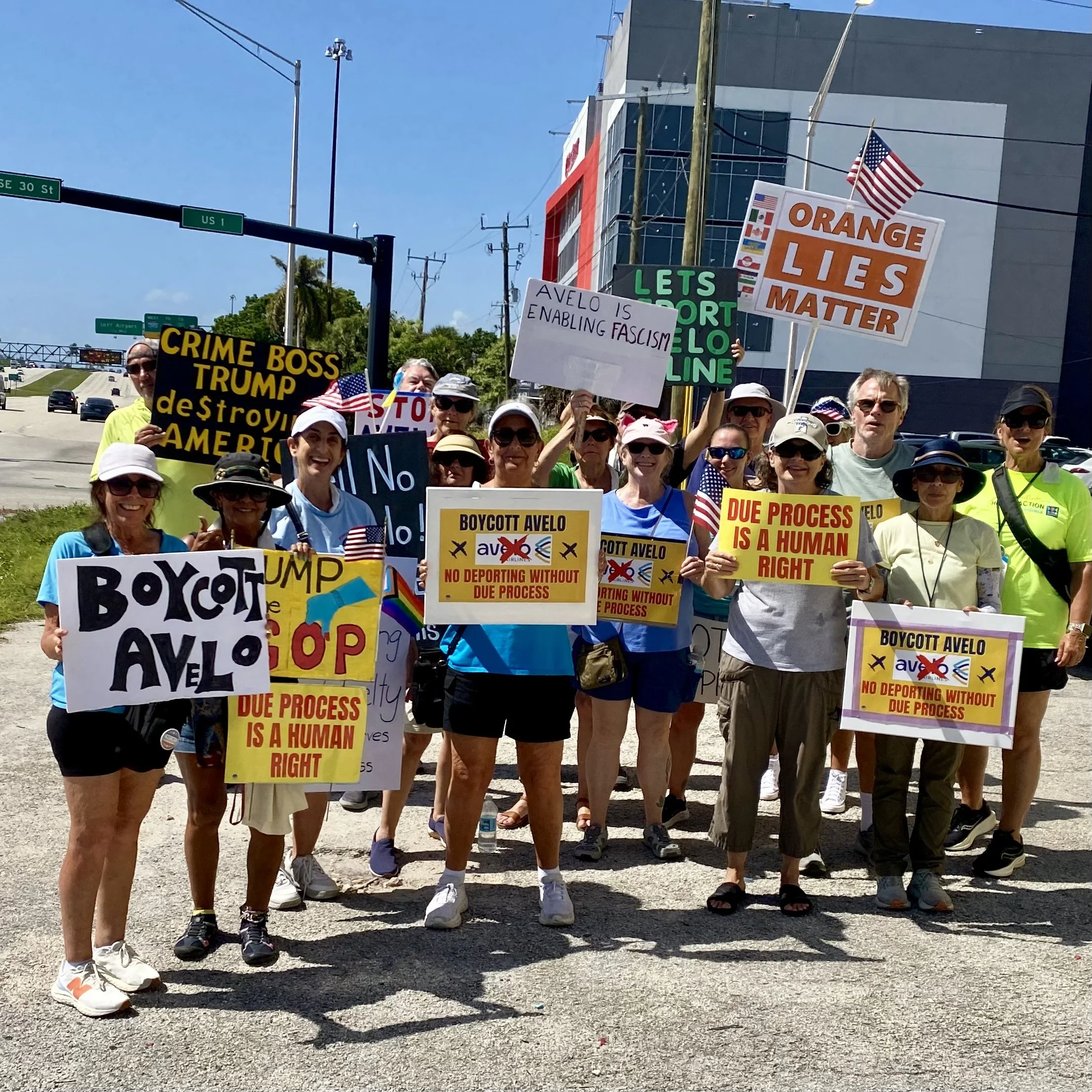 A group of protesters holding signs and American flags on a sunny day in an outdoor urban area. Signs include messages like 'Boycott AVELO,' 'Orange Lies Matter,' and 'Due Process is a Human Right.'
