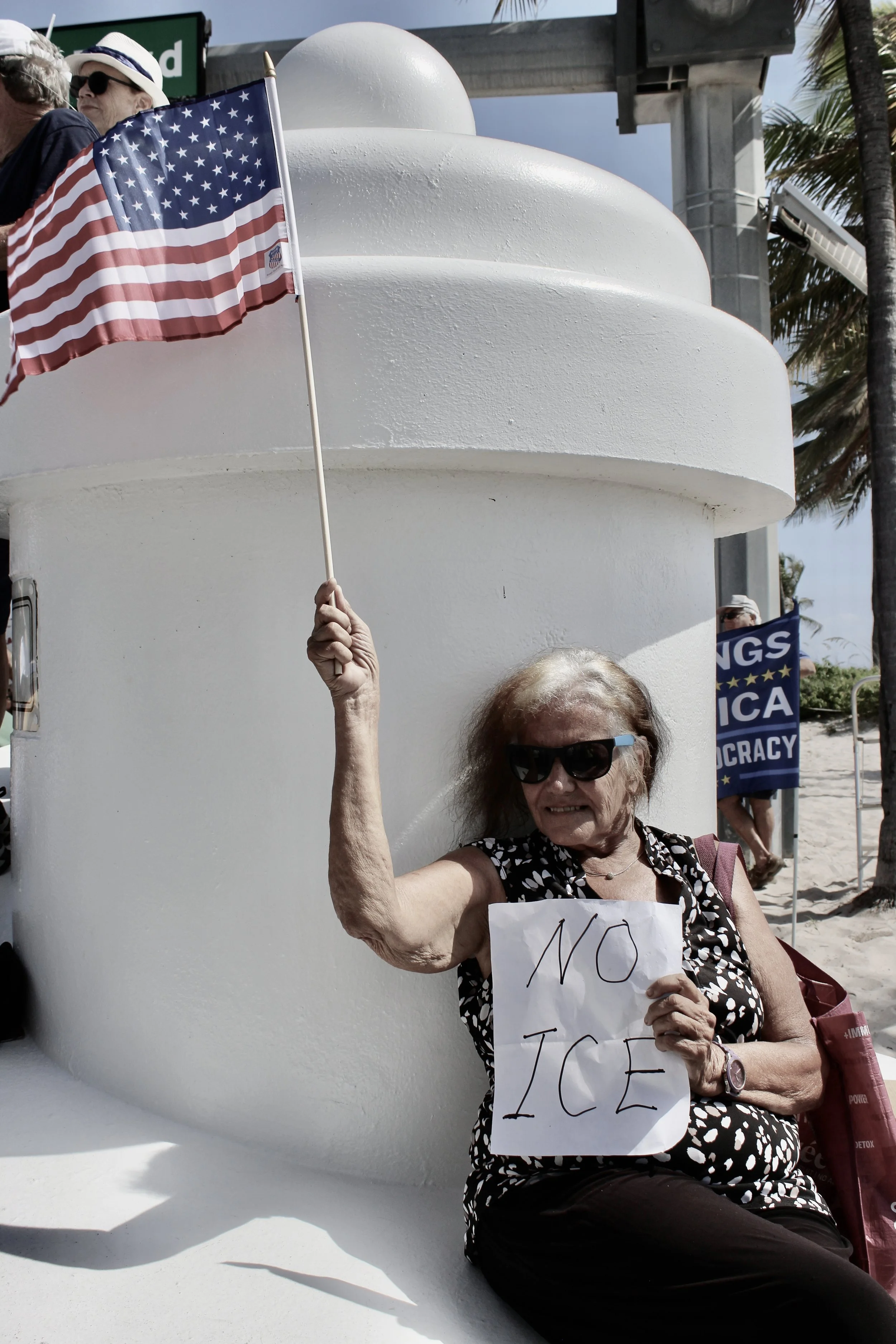 An elderly woman wearing sunglasses and a black and white patterned dress sitting outdoors at a protest, holding a sign that says 'NO ICE' and waving an American flag. She is seated beside a white structure, possibly part of a monument or building, with some protesters and banners visible in the background.