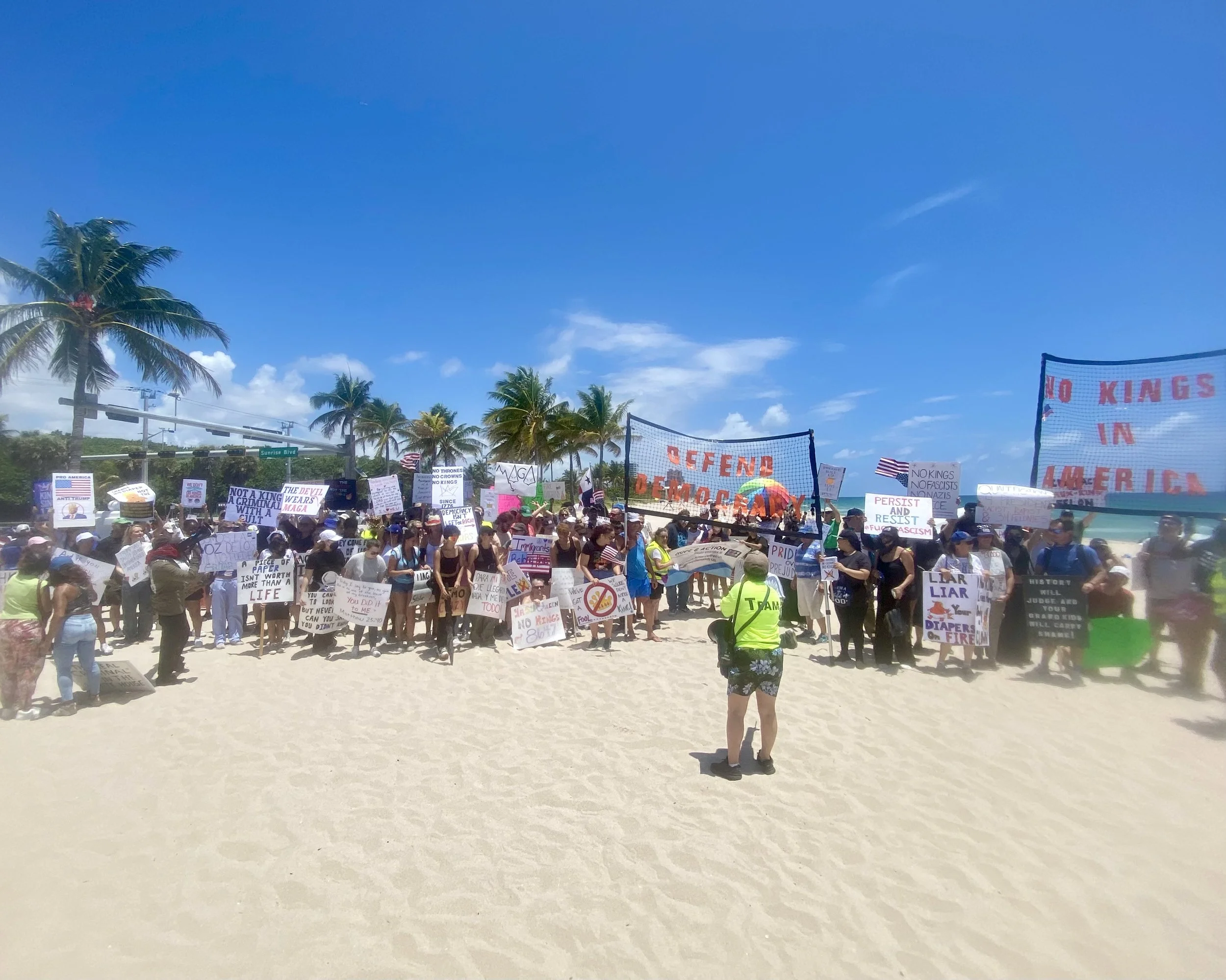 A large group of people gathered on a beach for a protest or rally, holding various signs and banners. There are palm trees and a bright, partly cloudy sky in the background.