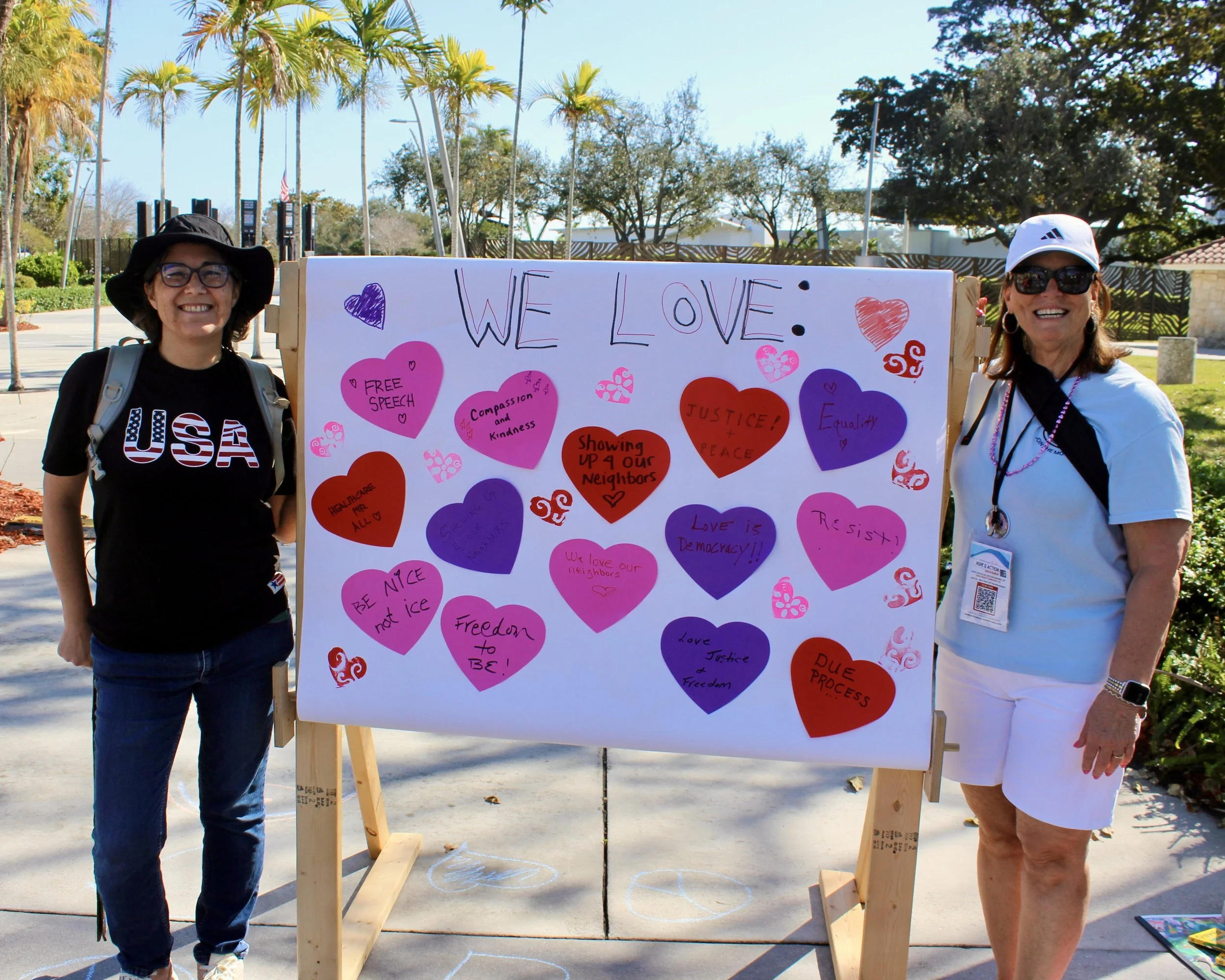 Two women standing on either side of a large white poster board decorated with text and multicolored heart cutouts. The poster reads 'WE LOVE' and includes messages such as 'Free Speech,' 'Compassion and Kindness,' 'Justice! Peace,' and 'Equality.' The women are outdoors, with trees and a fence in the background, and are smiling at the camera.