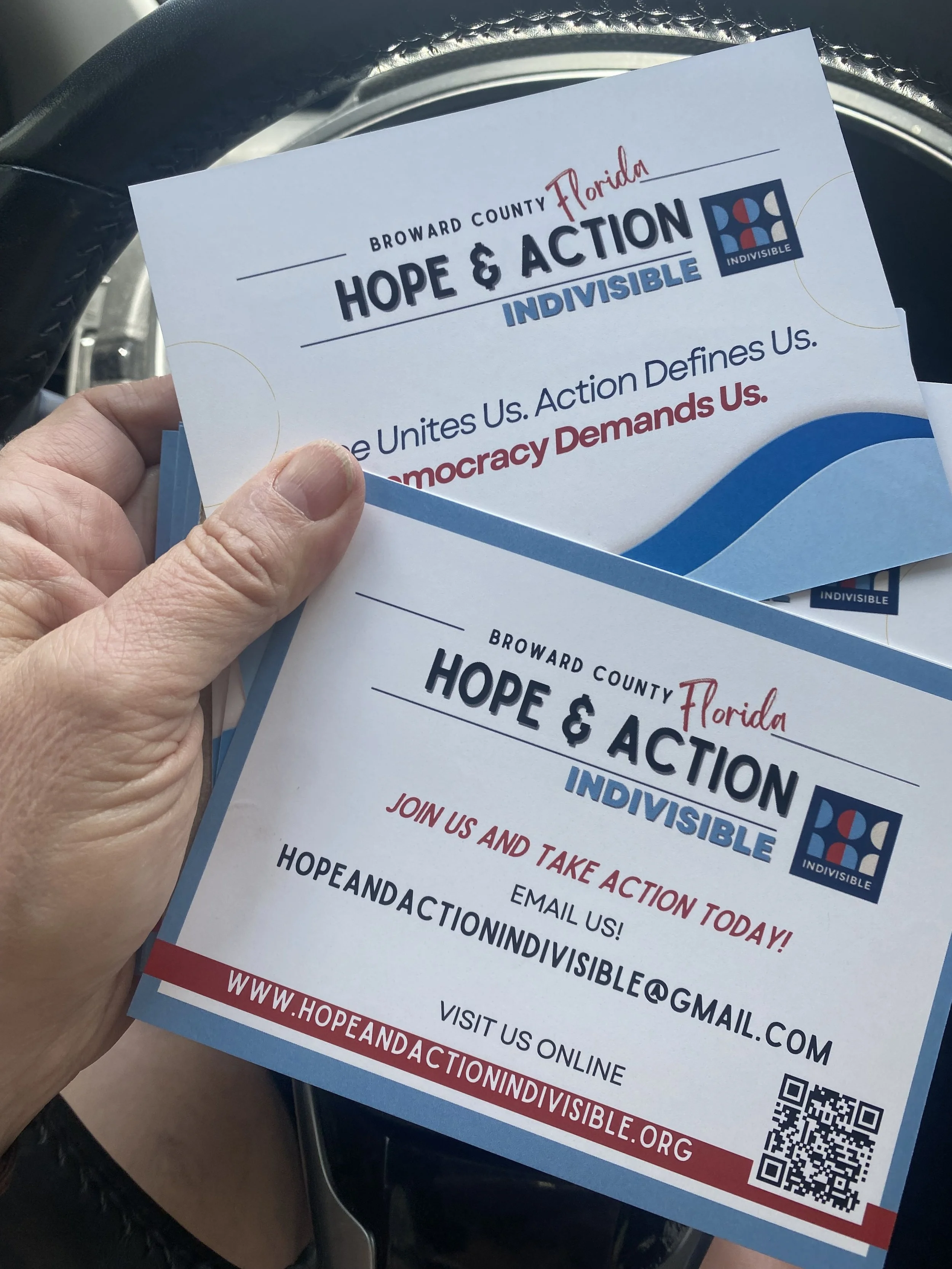 A hand holding flyers promoting Hope & Action, an initiative by Broward County, Florida. The flyers feature the words 'Hope & Action Indivisible' and encourage joining and taking action today. The flyer includes a website, email, and QR code for more information.