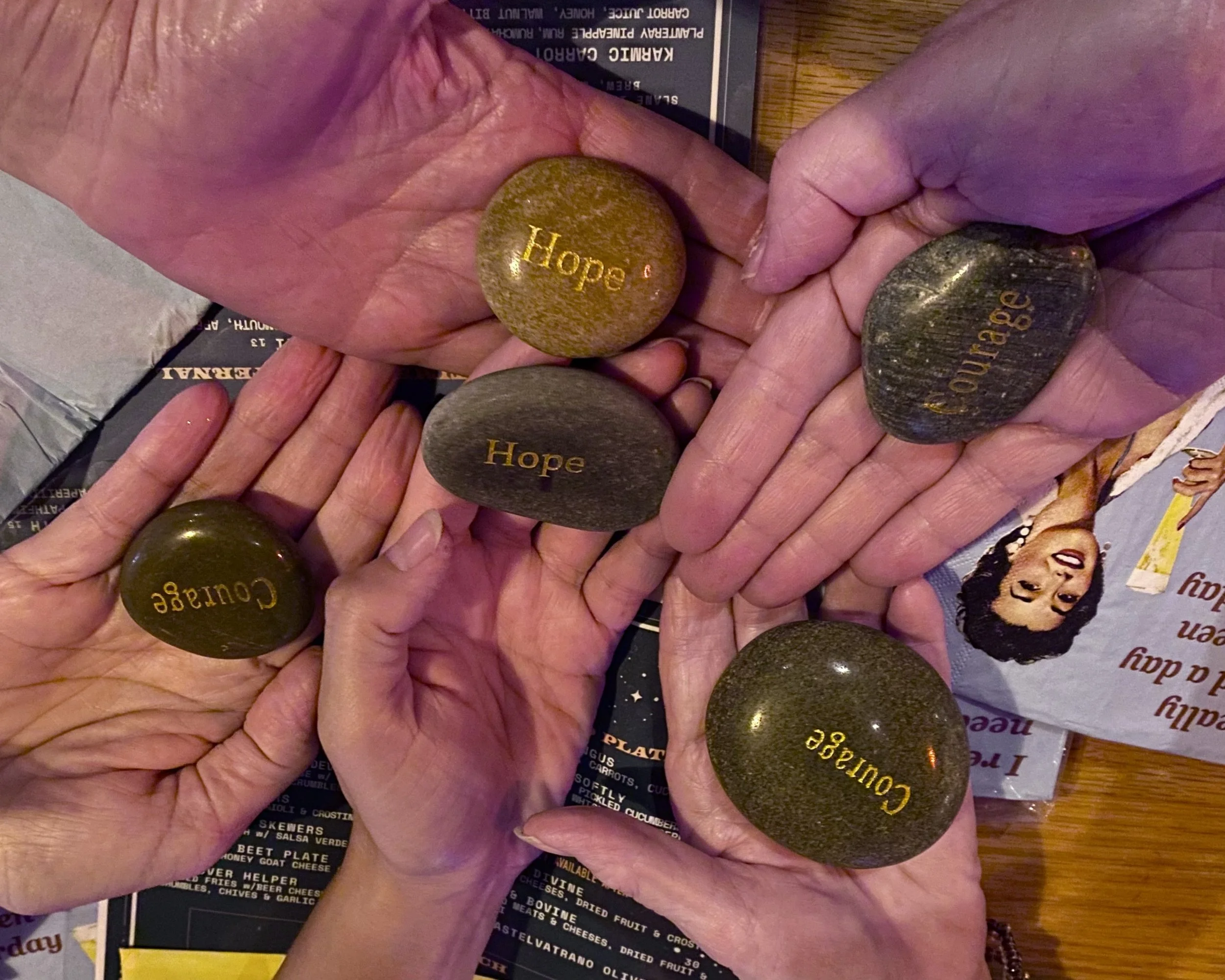 Close-up of multiple stones with a smooth surface, engraved with the words 'Hope,' 'Courage,' and 'Acceptance,' held by hands over a menu or magazine.
