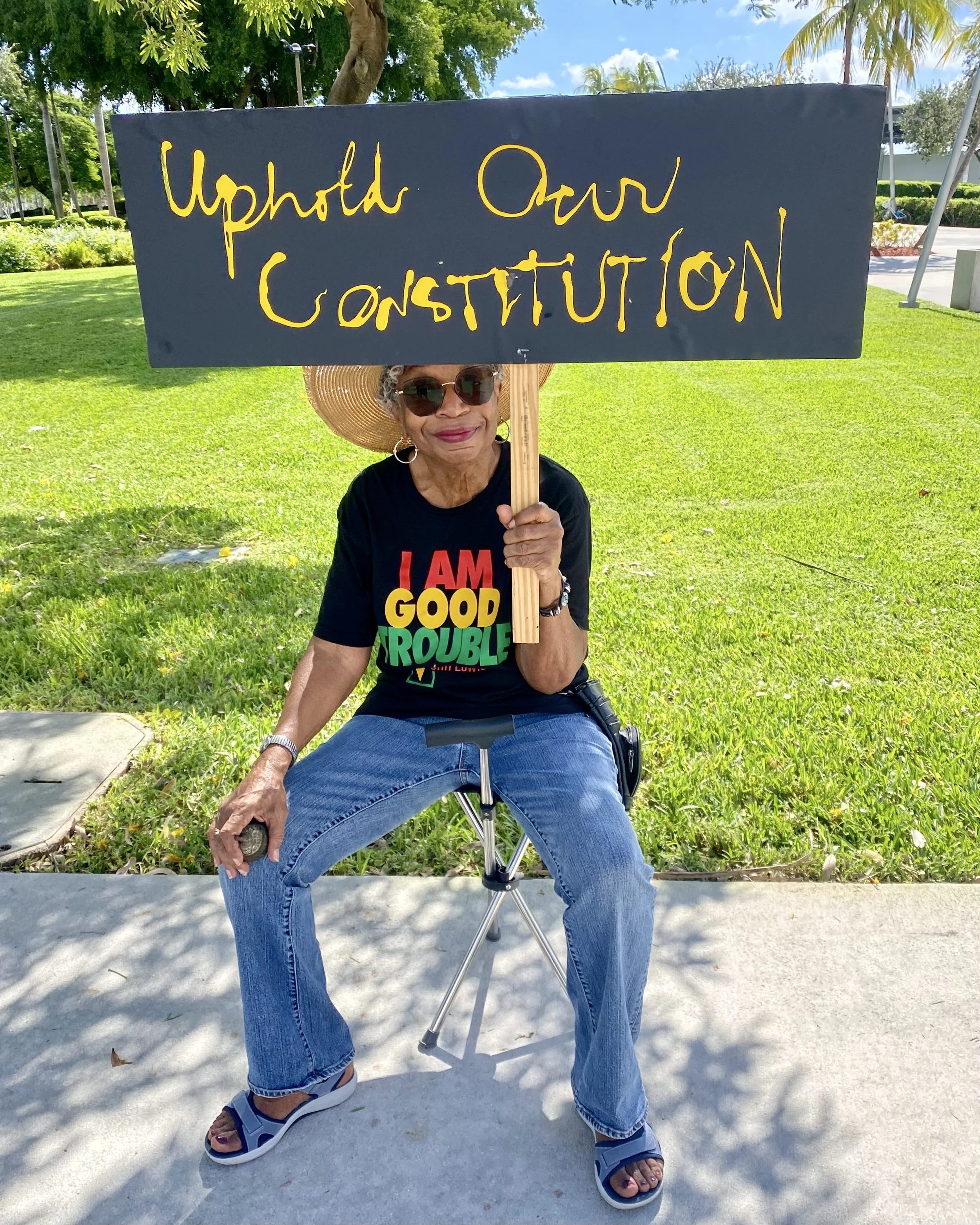 An elderly woman sitting on a foldable chair outdoors on a sunny day, holding a sign that says 'Upvote Our CONSTITUTION' with her right hand. She is wearing sunglasses, a large sunhat, a black t-shirt with colorful text, blue jeans, and sandals. The background features green grass, trees, and a clear blue sky.