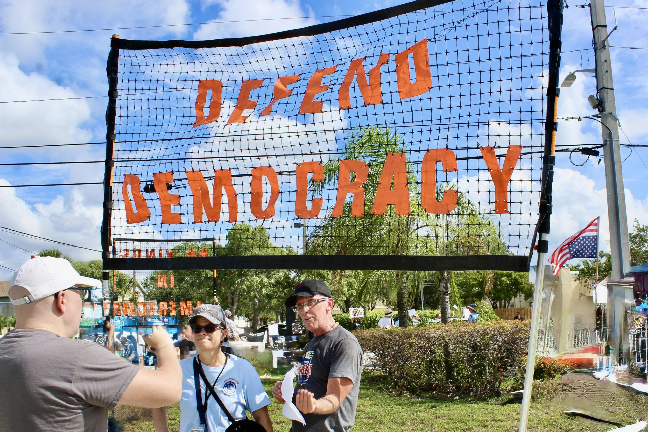 People gathered outdoors at a festival, standing under a sign that reads 'Defend Democracy' in orange letters, with tents and flags visible in the background.
