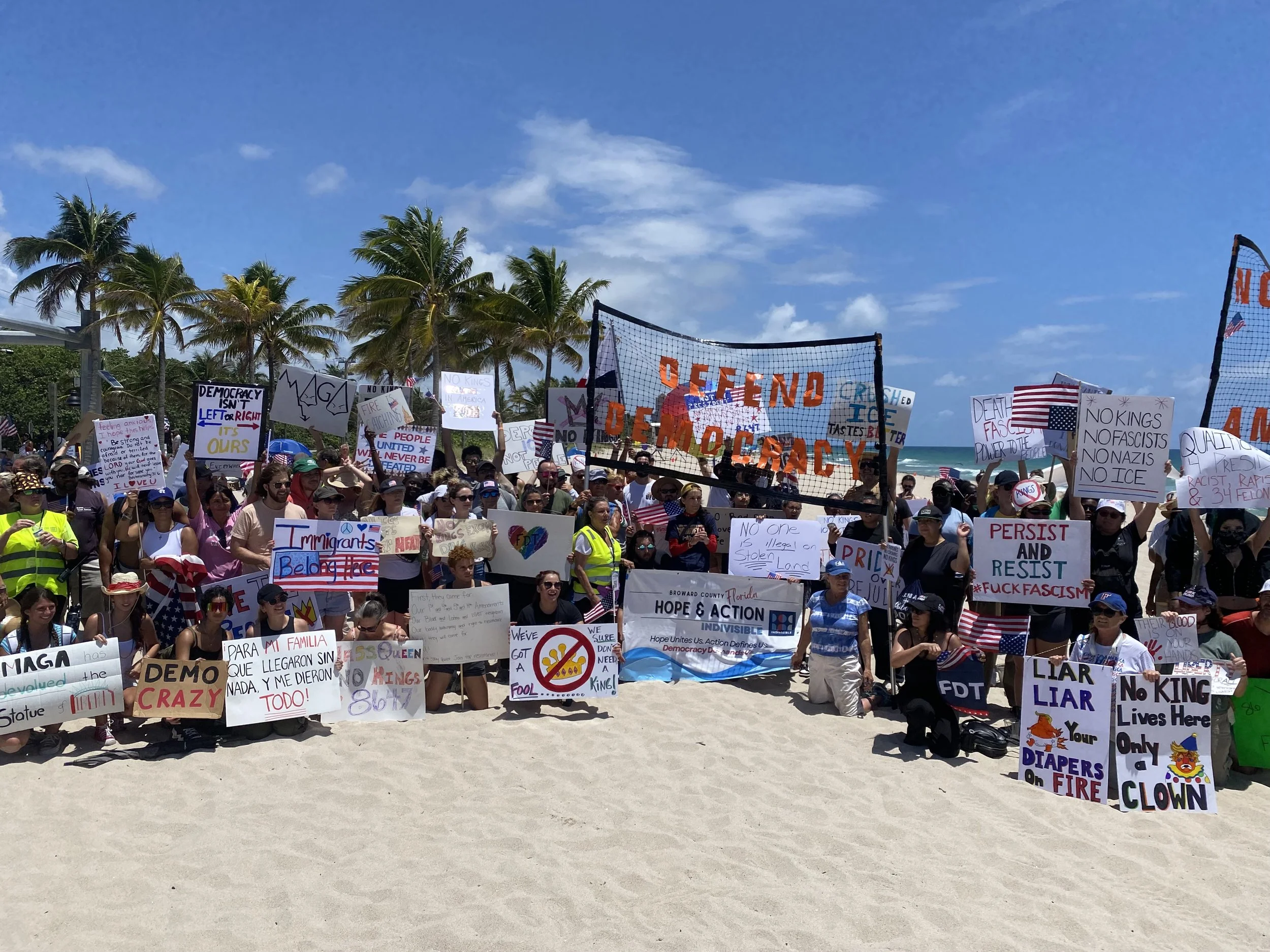 A large crowd of people participating in a protest or demonstration on a beach, holding signs with messages about democracy, immigration, and political issues. Palm trees and a blue sky are visible in the background.