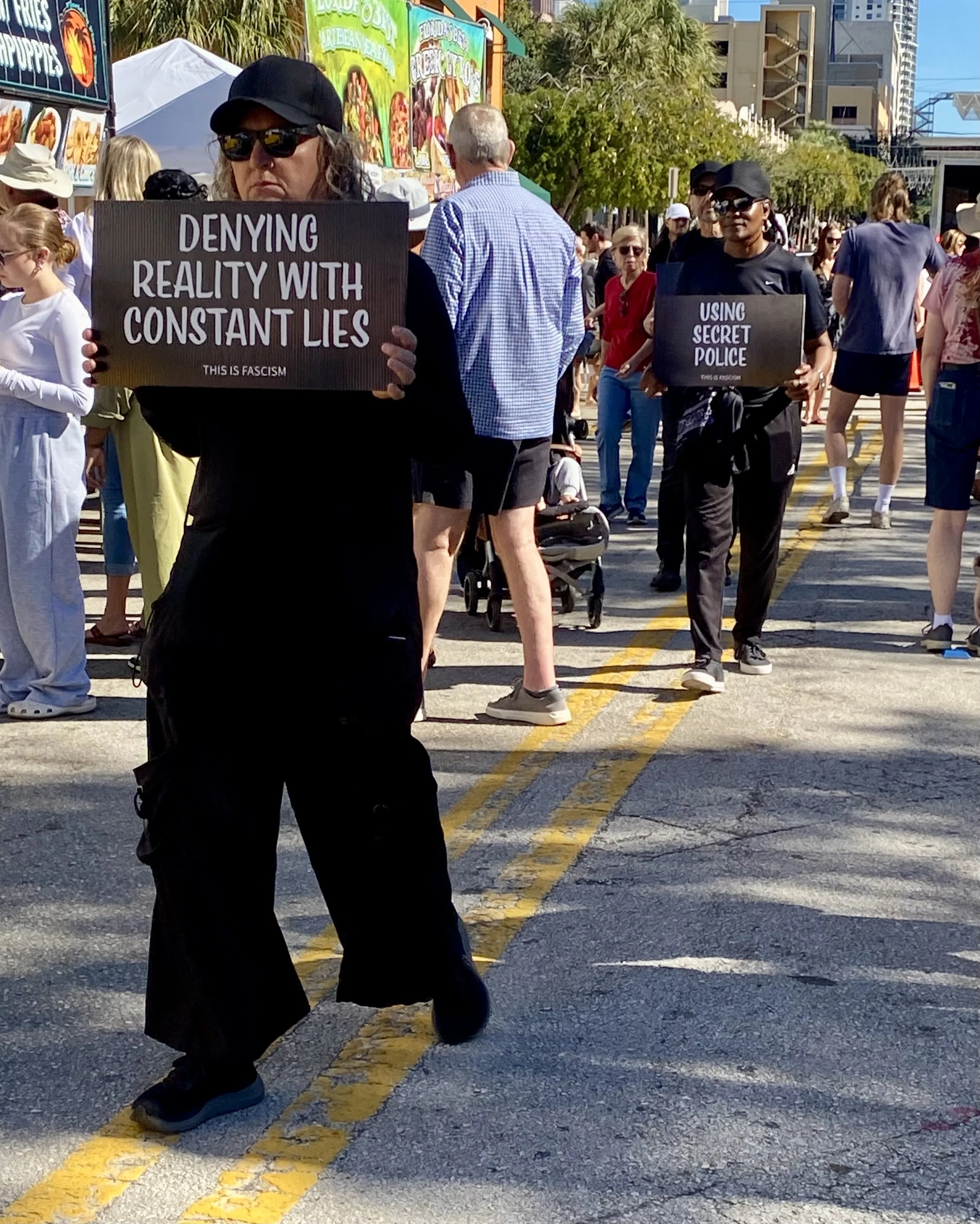 A group of people walking in a street protest or demonstration. Several individuals are holding signs with messages, and there are food stalls and trees in the background.