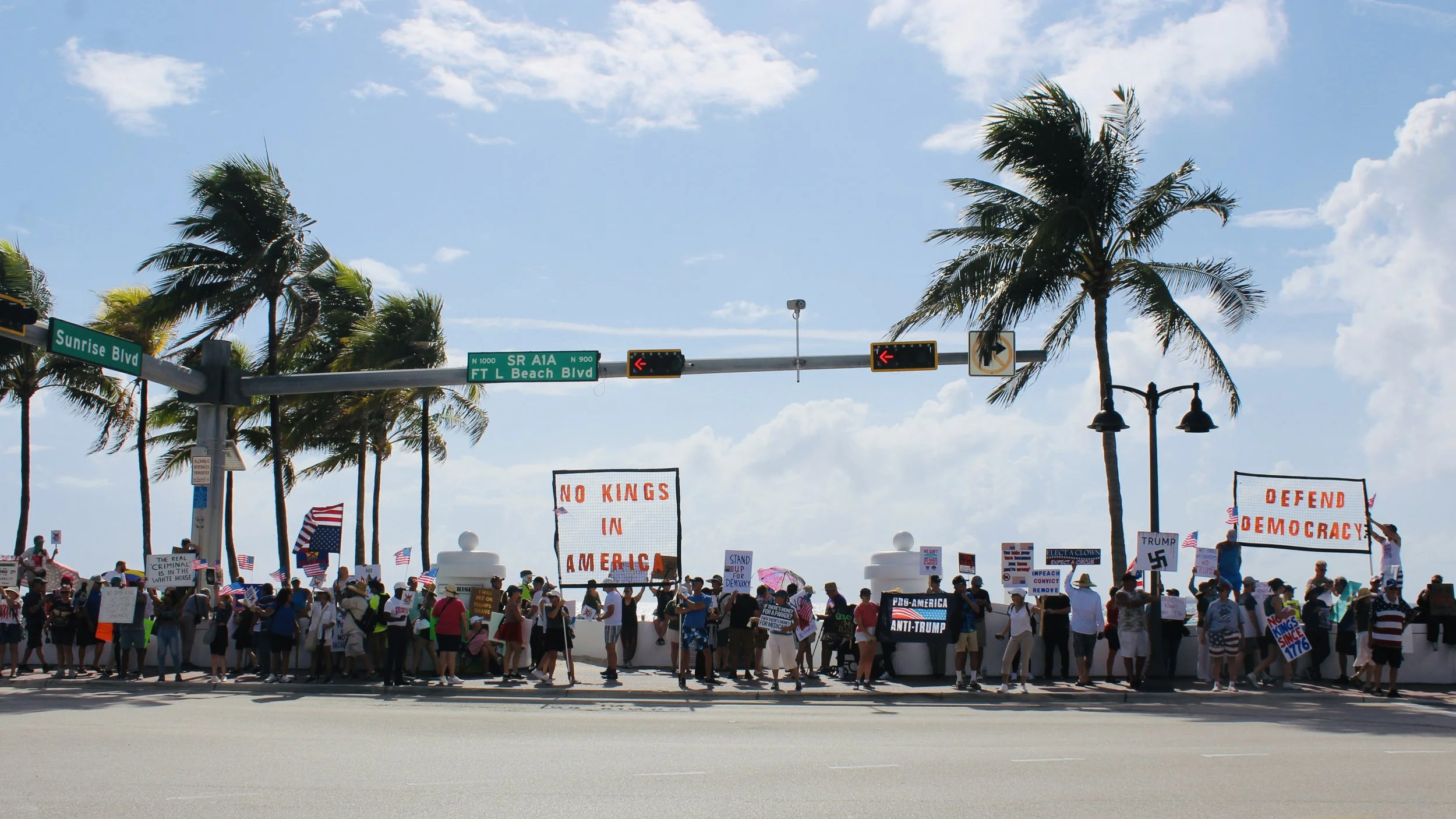 A large crowd protesting at a rally with signs and American flags, with palm trees, street signs, and a blue sky in the background.